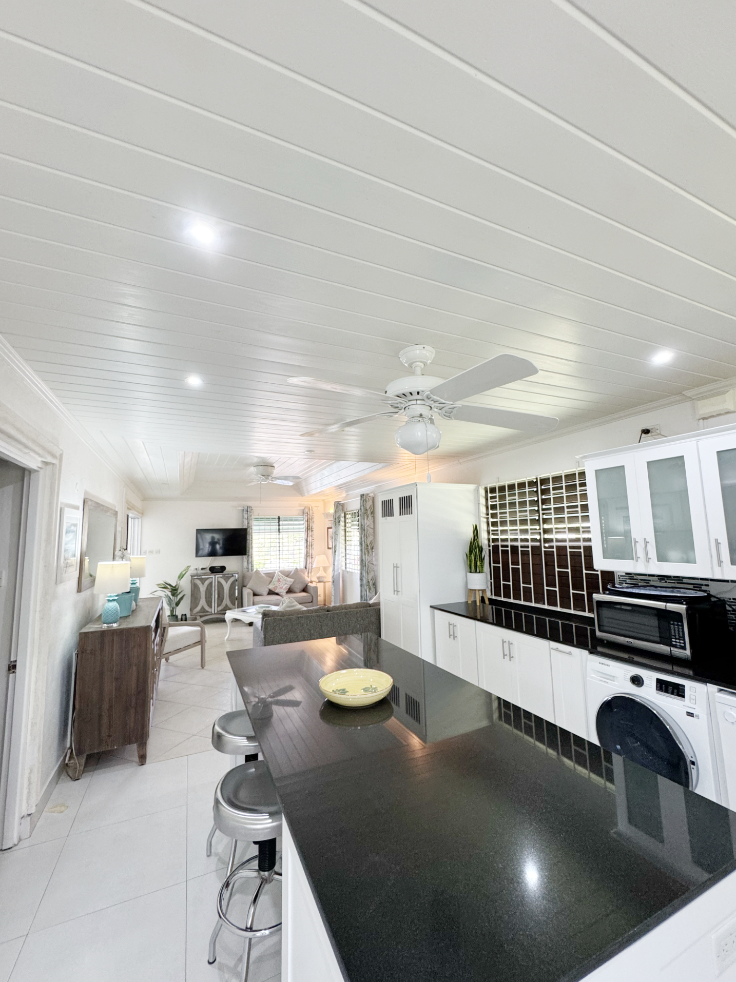 Interior view of a living room and kitchen area with white cabinetry, black countertop, ceiling fan, and a dining table with stools.