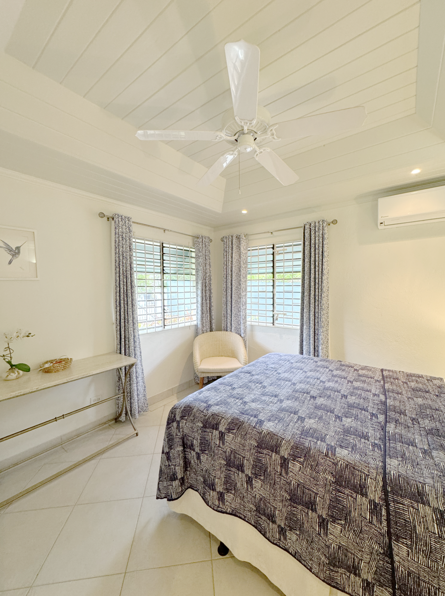 A bedroom with a bed covered by a patterned bedspread, a white armchair near the windows, a narrow table with decorative items on the left, three windows with patterned curtains, and a ceiling fan.