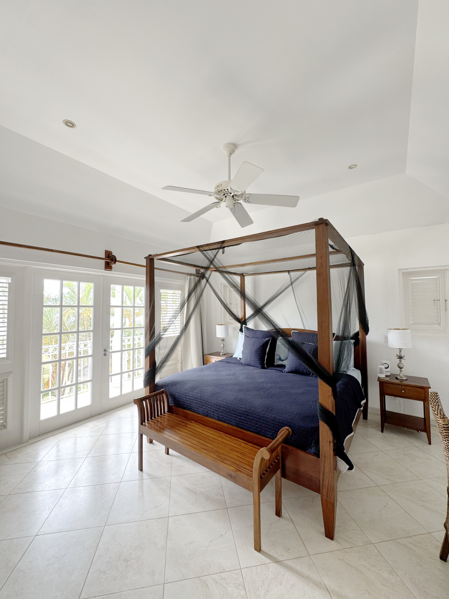 A bedroom with a four-poster bed draped with black netting, navy bedding, and matching pillows, with a wooden bench at the foot of the bed, bedside tables with lamps, and glass-paned doors leading outside.