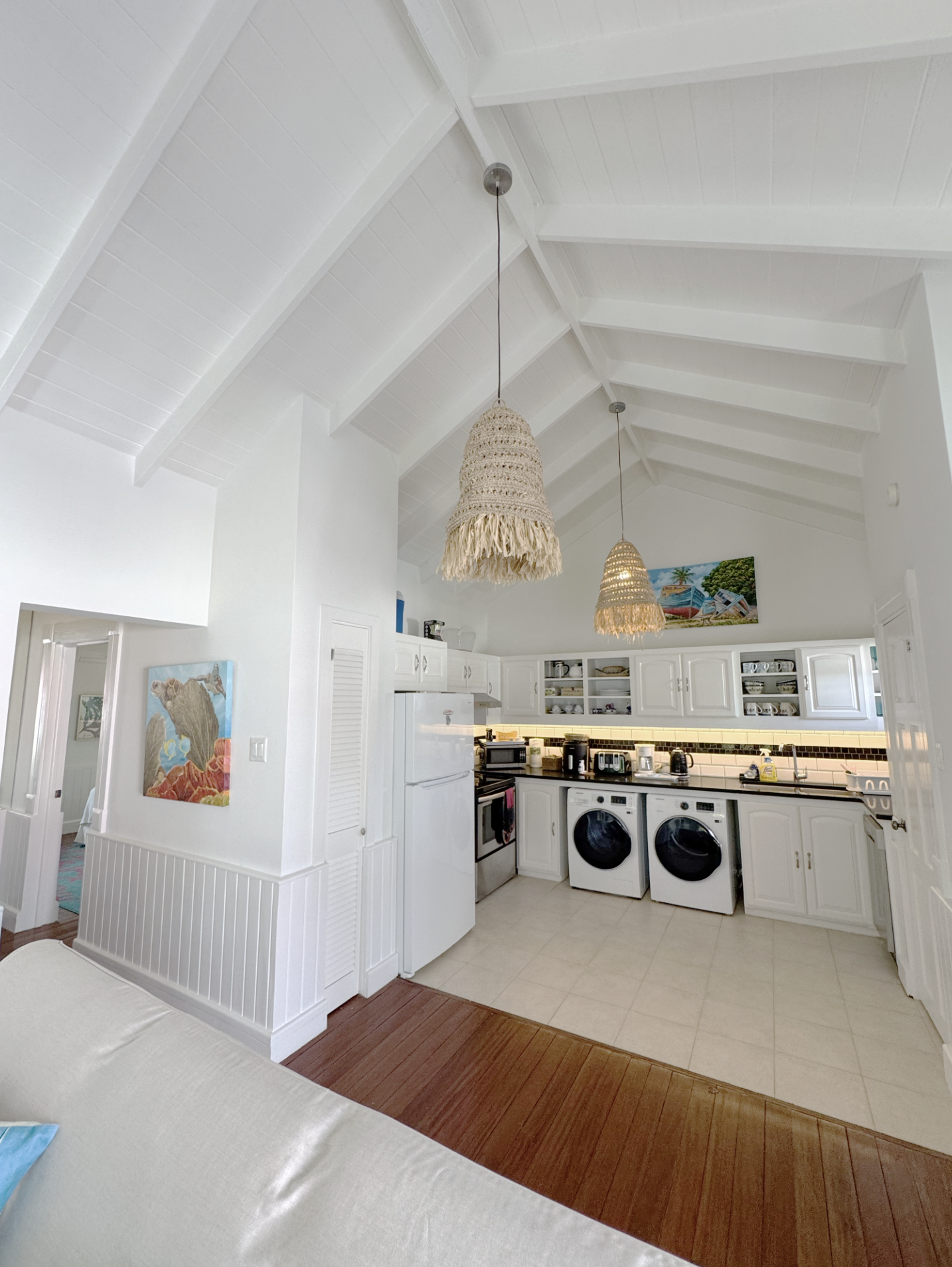A kitchen with white cabinets, a white refrigerator, and a countertop with various small appliances. Hanging from the ceiling are two woven pendant lights. On the wall, there's a colorful beach-themed painting. The floor is a combination of wooden an