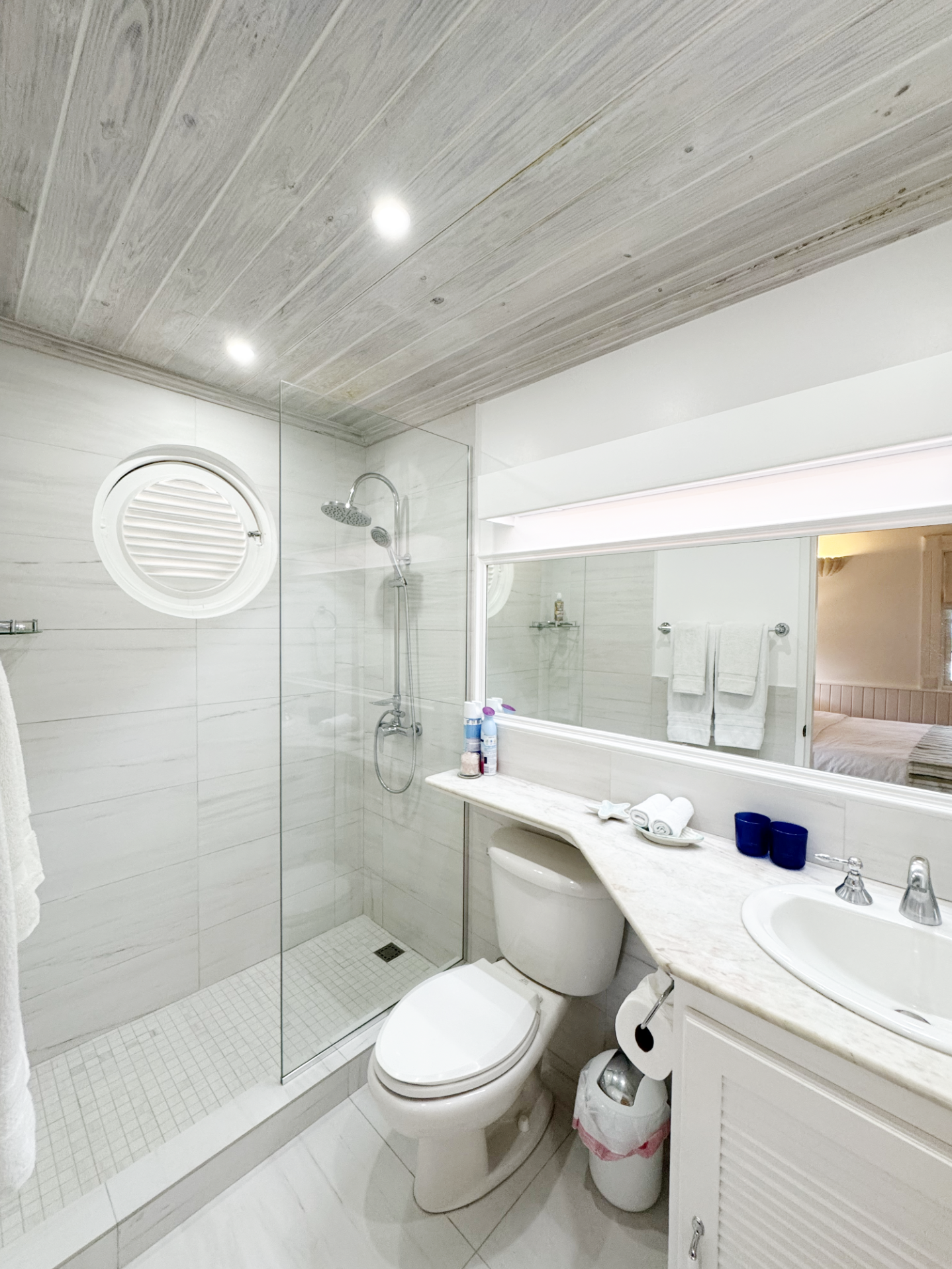 A modern bathroom with a glass-enclosed walk-in shower, a toilet, a white vanity with a marble countertop, a large mirror, and a bedroom visible through the door. The ceiling has wood paneling and recessed lighting.