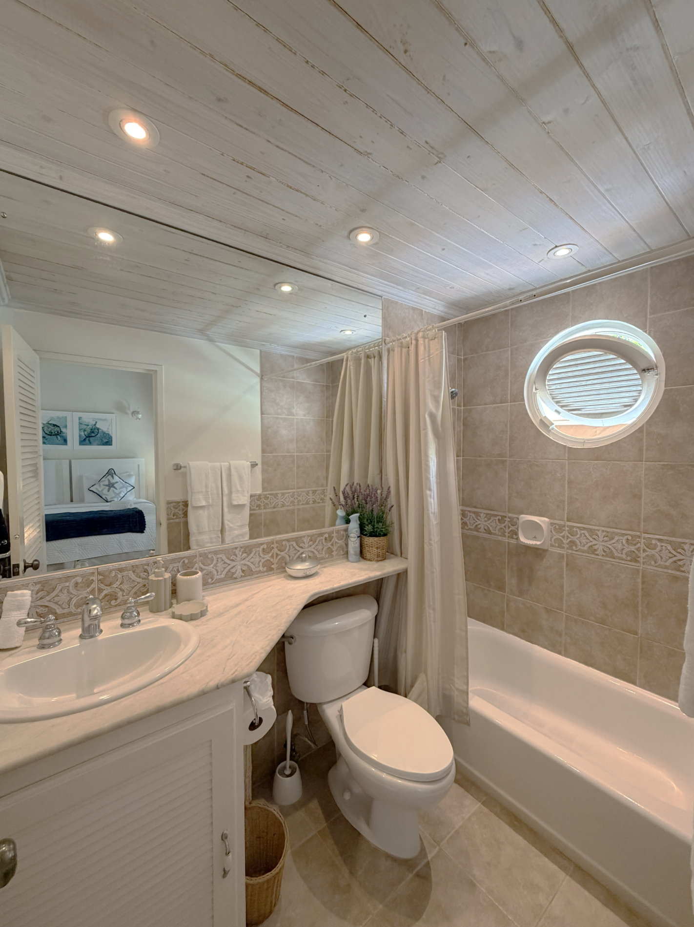 A bathroom with a white vanity, marble countertop, and a sink with a silver faucet. There's a toilet and a bathtub with a shower curtain. The wall tiles are beige with decorative border tiles. An oval window lets in natural light. The ceiling is whit
