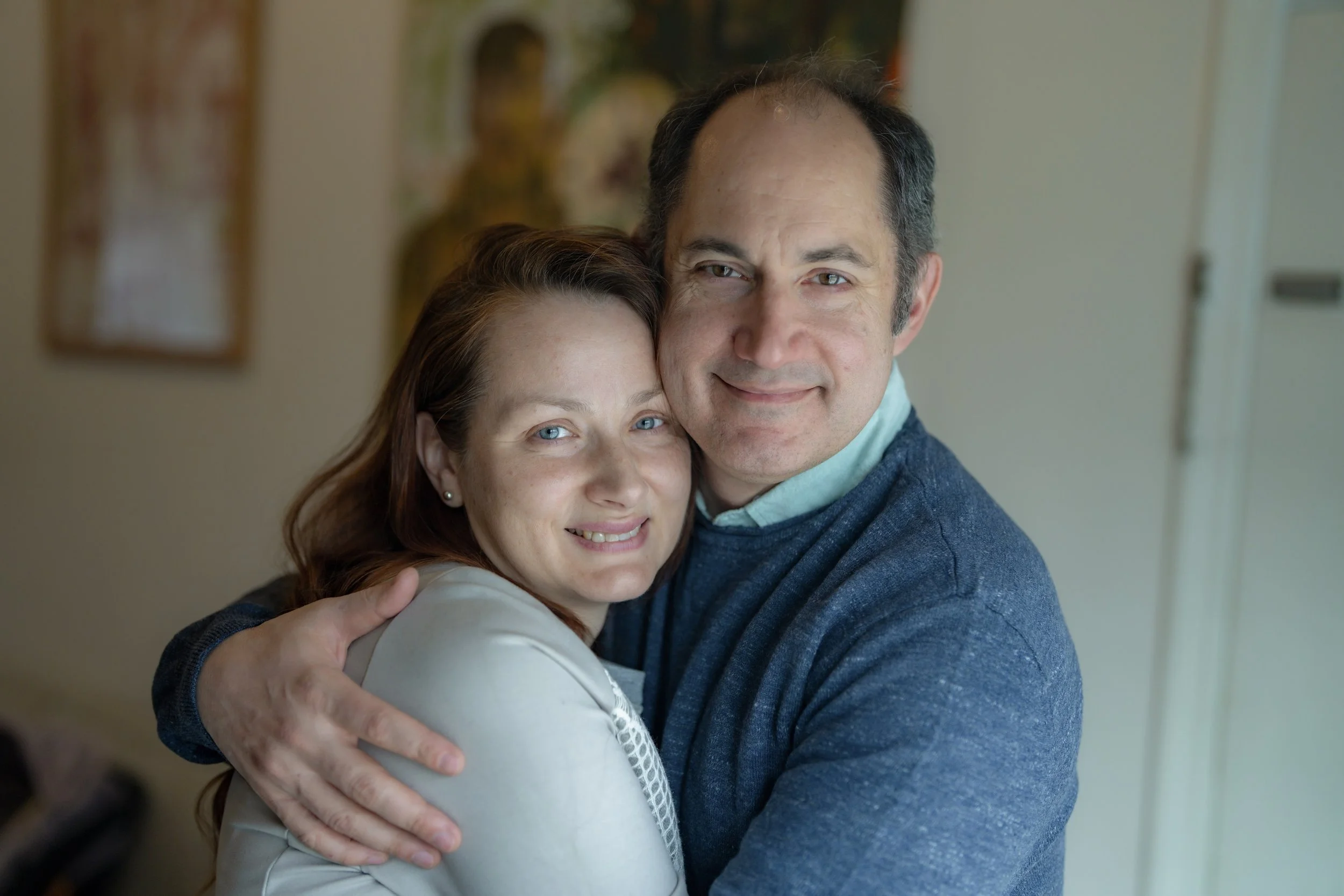 Bonenberger and his wife hugging each other, smiling at the camera. Iryna  has reddish-brown hair and is wearing a light-colored jacket. Adrian has dark hair graying at the temples and is wearing a blue sweater over a light blue collared shirt.