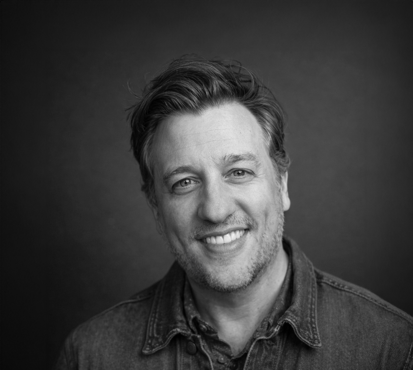 Black and white portrait of a smiling man with short, wavy hair, wearing a denim jacket.