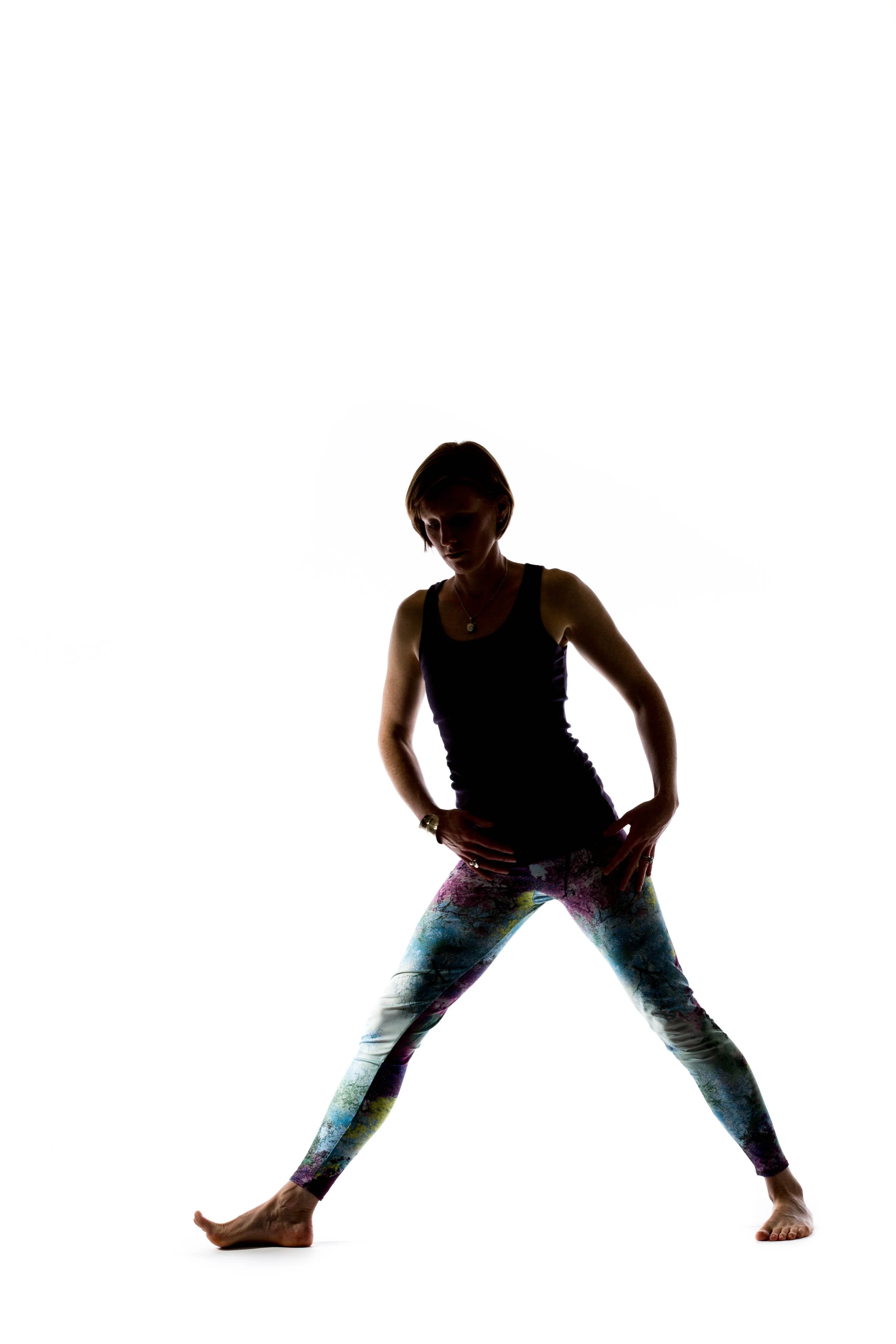 Esther Arrowsmith yoga teacher and creator of SLEEPWELL RITUALS. standing in a wide stance with her hands on her hips, wearing colorful patterned leggings and a black tank top, against a white background.