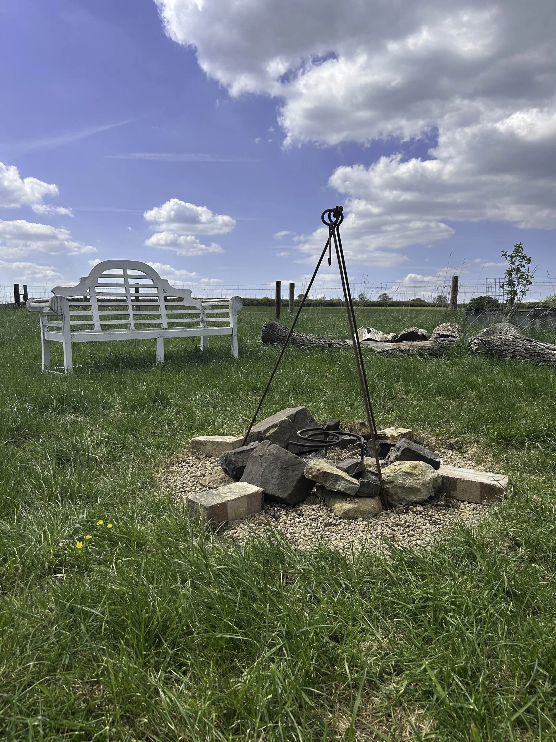 A fire pit with a metal tripod over bricks and stones, situated on grassy land with a white wooden bench in the background under a partly cloudy sky.
