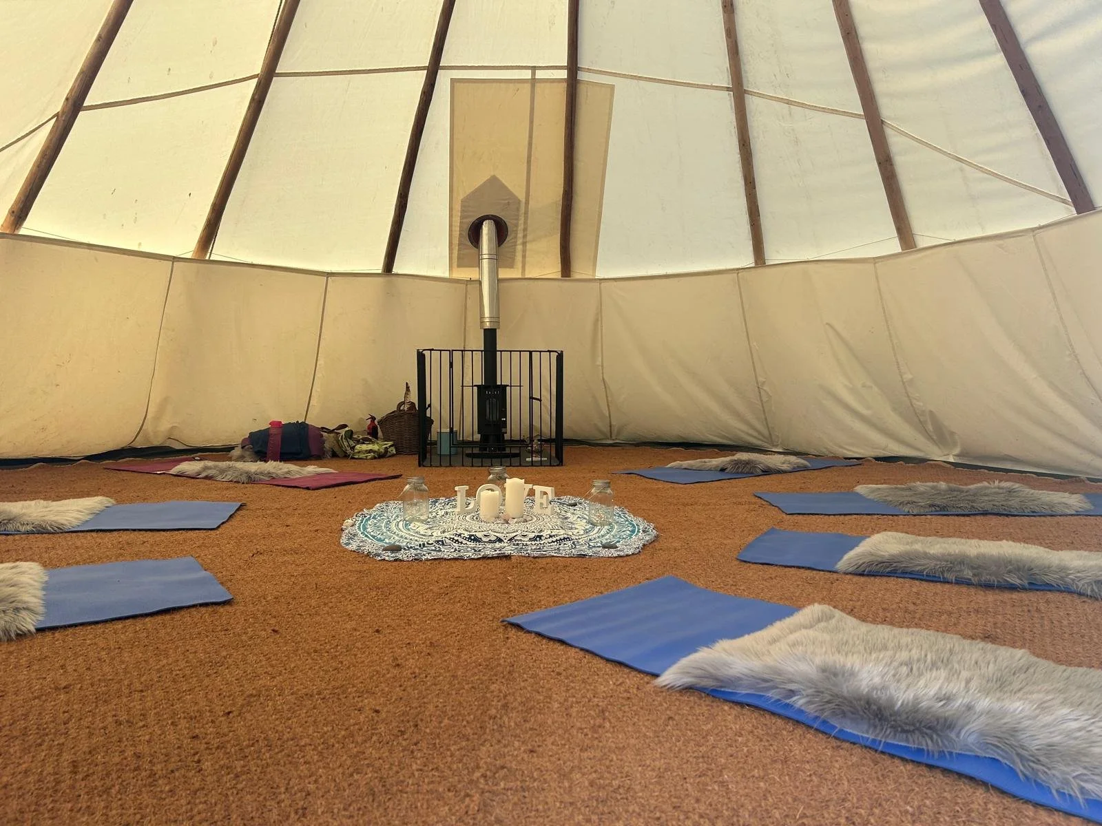 Interior of a tipi with a small stove at the center, surrounded by blue yoga mats with fur rugs, candles, and pillows for meditation or relaxation.