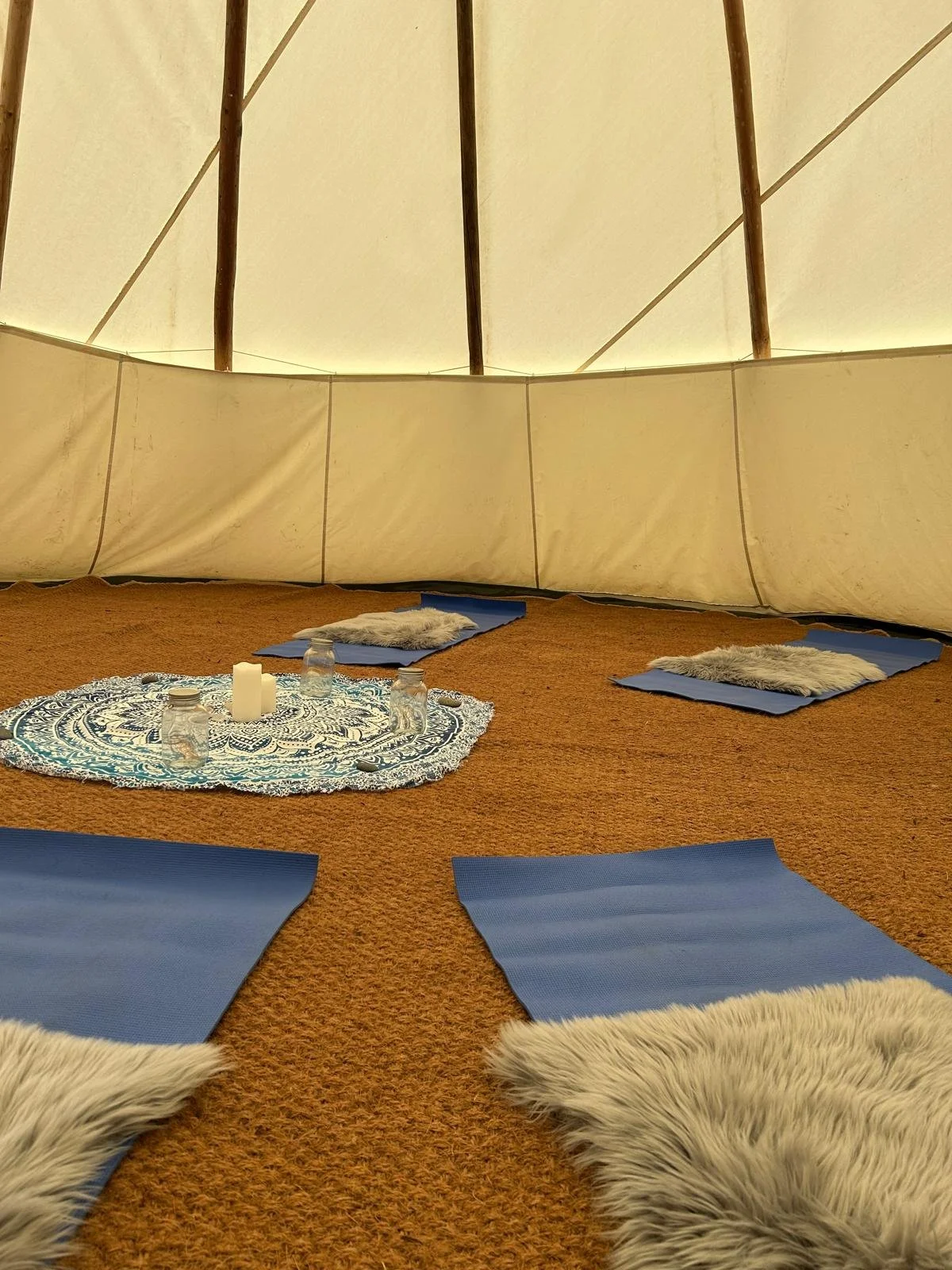 Interior of a tipi with blue yoga mats and faux fur rugs arranged around a central round cloth with candles and jars, on a brown carpet.