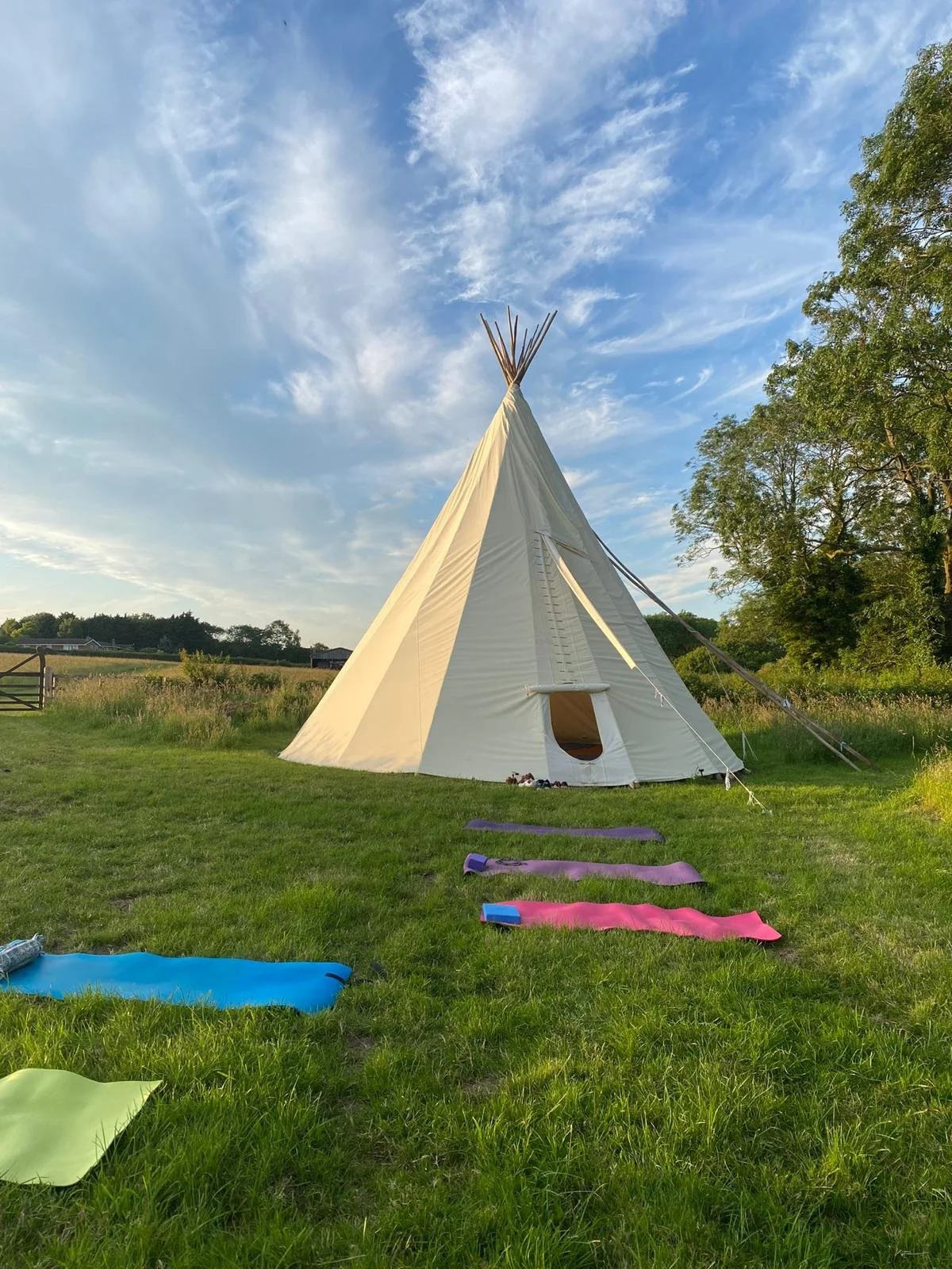 A white tipi tent set up on a grassy field with yoga mats lined up in front, trees in the background, and a partly cloudy sky overhead.