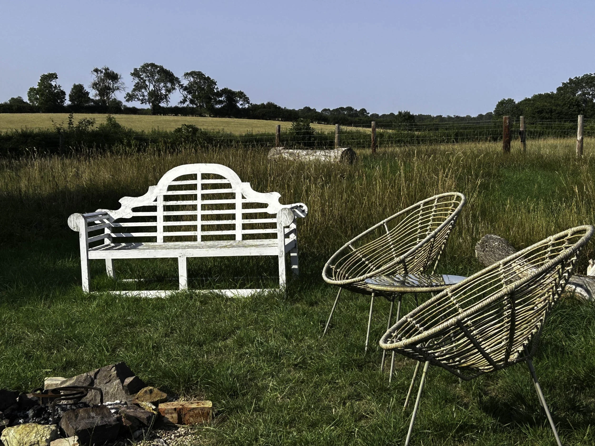 Outdoor scene with a white wooden bench, two rattan chairs, and a campfire surrounded by rocks on a grassy field. In the background, there is a barbed wire fence and a landscape of trees and rolling hills under a blue sky.