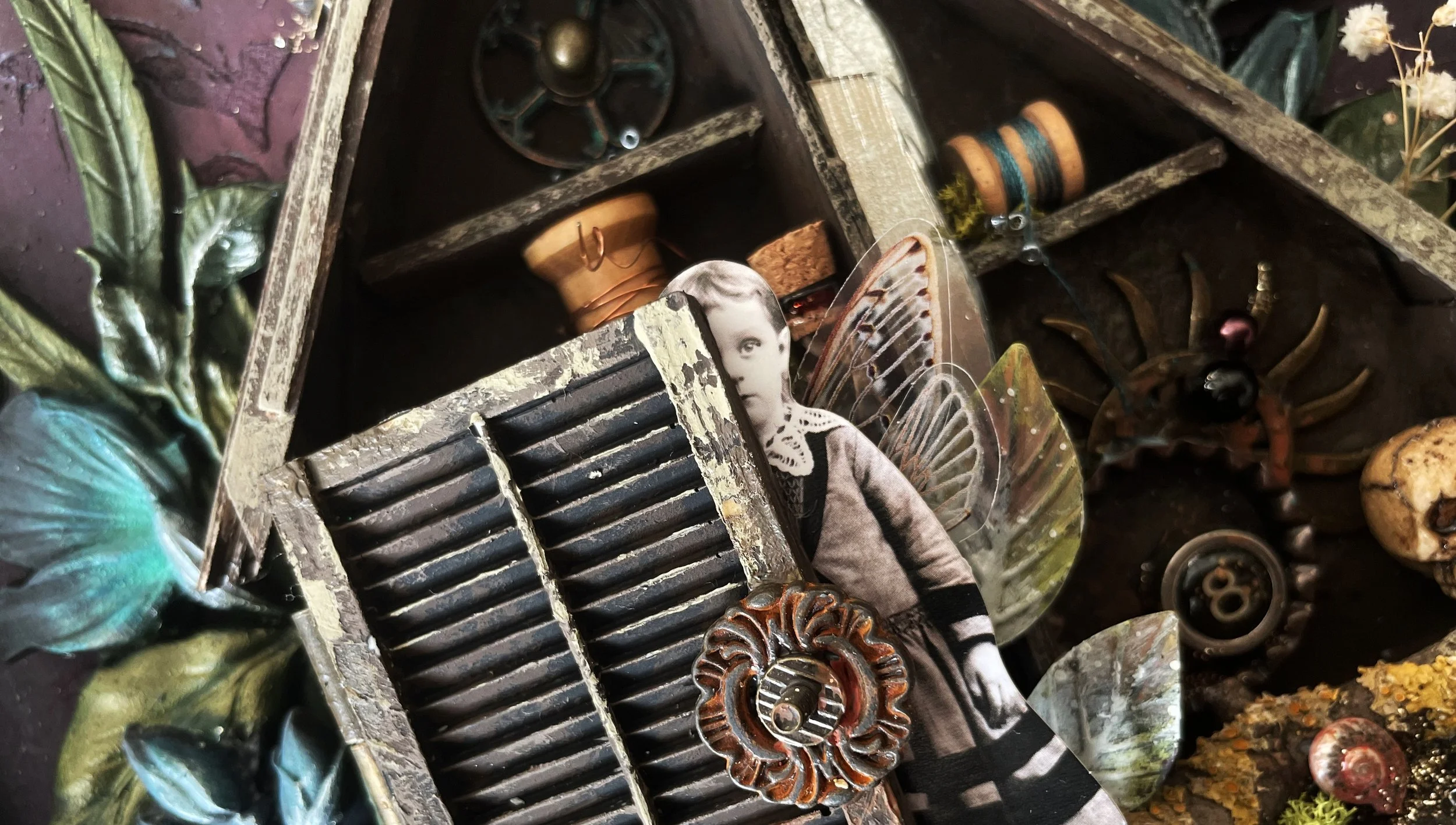 Close-up of a weathered, vintage wooden box filled with various small objects, including a black-and-white cutout of a child's face, a decorative butterfly wing, a metal gear, a tiny skull, and several spools of thread. The box is surrounded by a textured surface with painted leaves in green and blue hues.