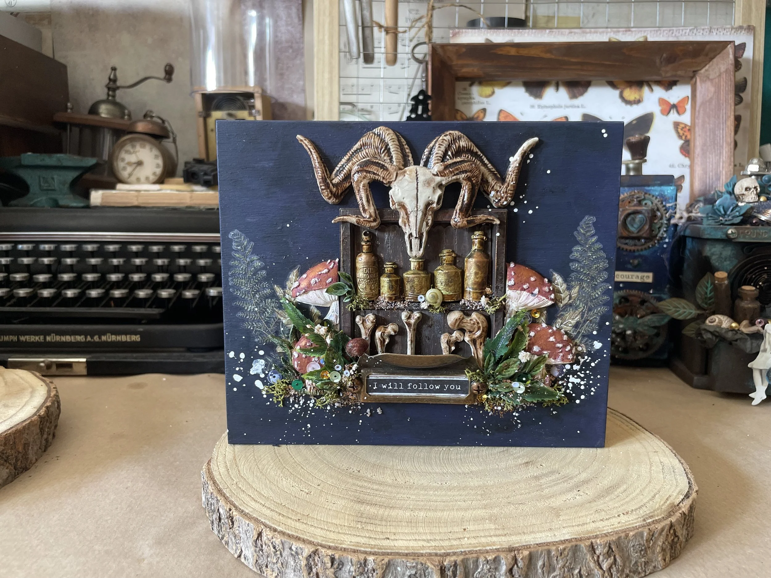 Decorative art piece with a ram skull, mushrooms, plants, and bottles on a dark background, with the phrase "I will follow you" at the bottom.