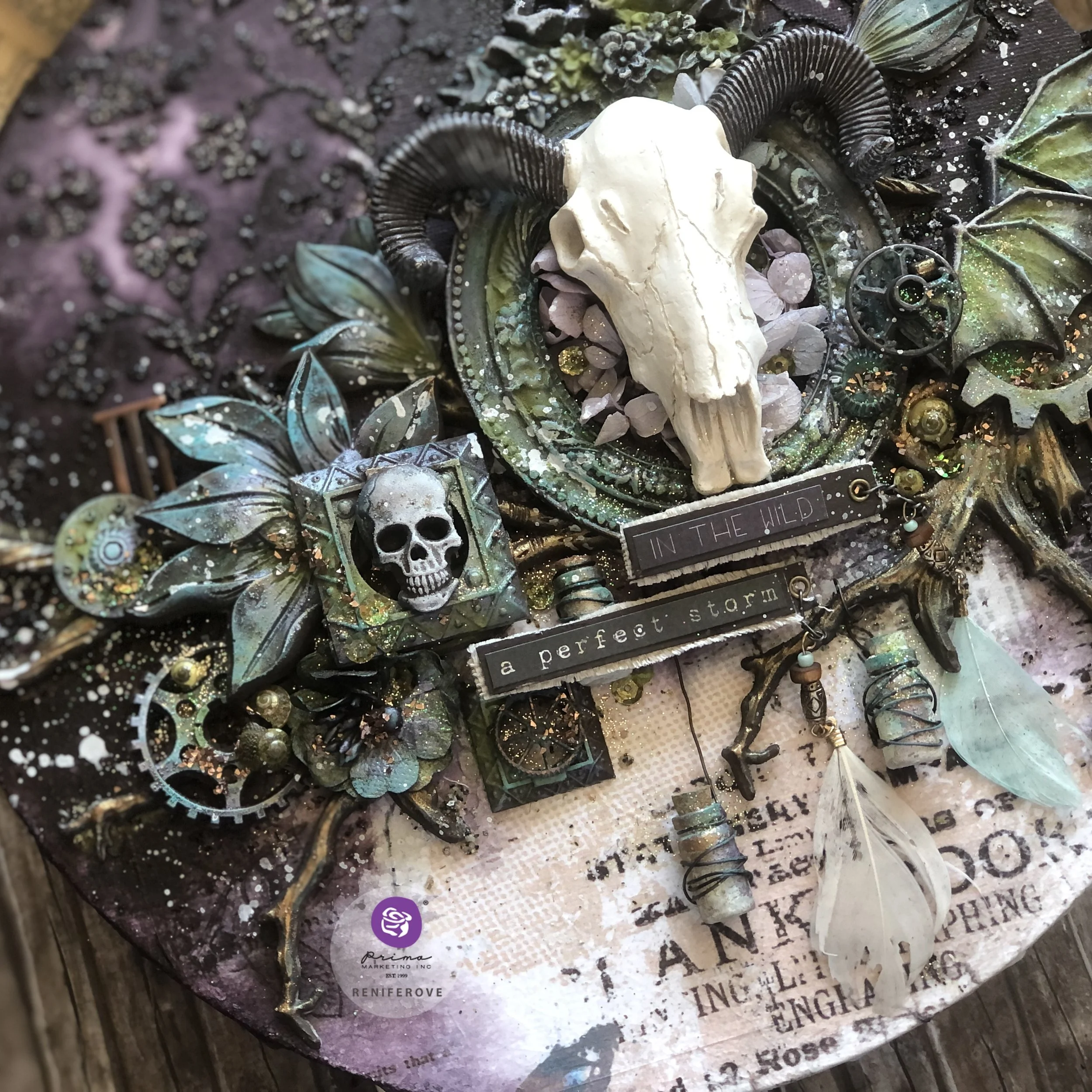 Mixed media art piece with skull and animal skull, feathers, gears, flowers, and decorative elements in a dark, earthy theme with text reading 'In the wild, a perfect storm.'