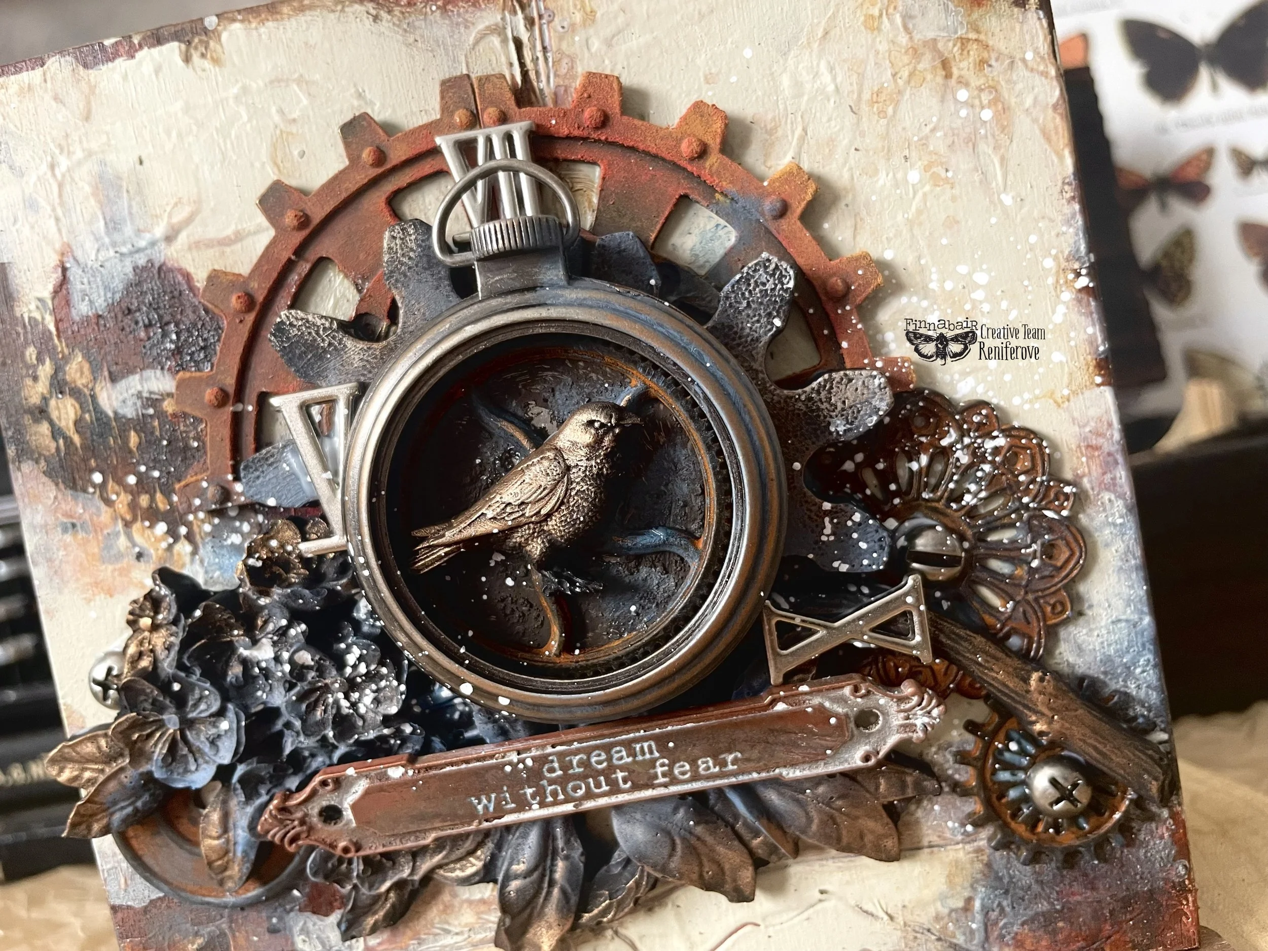 Mixed media artwork featuring a bird figurine inside a gear, metallic flowers, a metal banner reading "dream without fear," and butterfly and floral accents in brown, bronze, and silver tones.