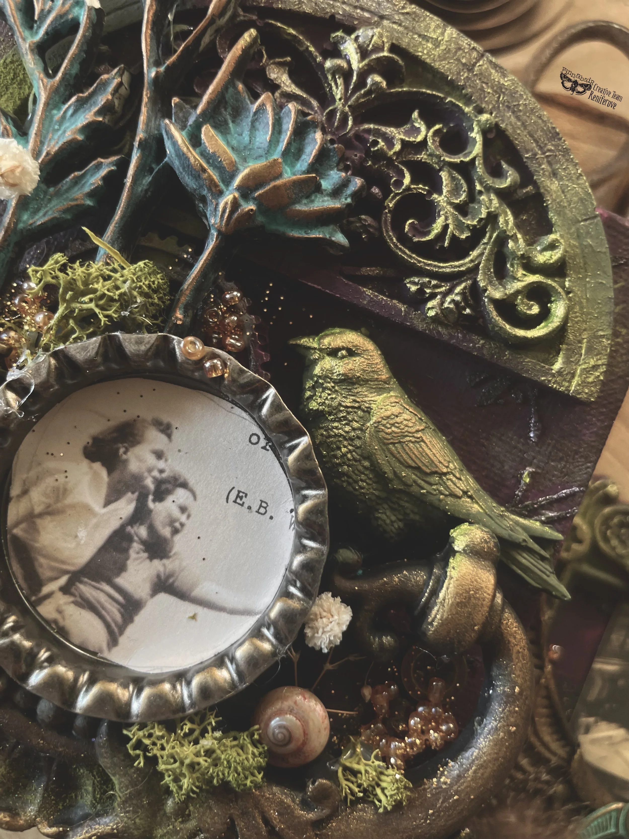 A decorative display featuring vintage items, including a black and white photograph of two women, ornate metalwork, a small bird sculpture, seashells, moss, beads, and an intricately designed metal piece.