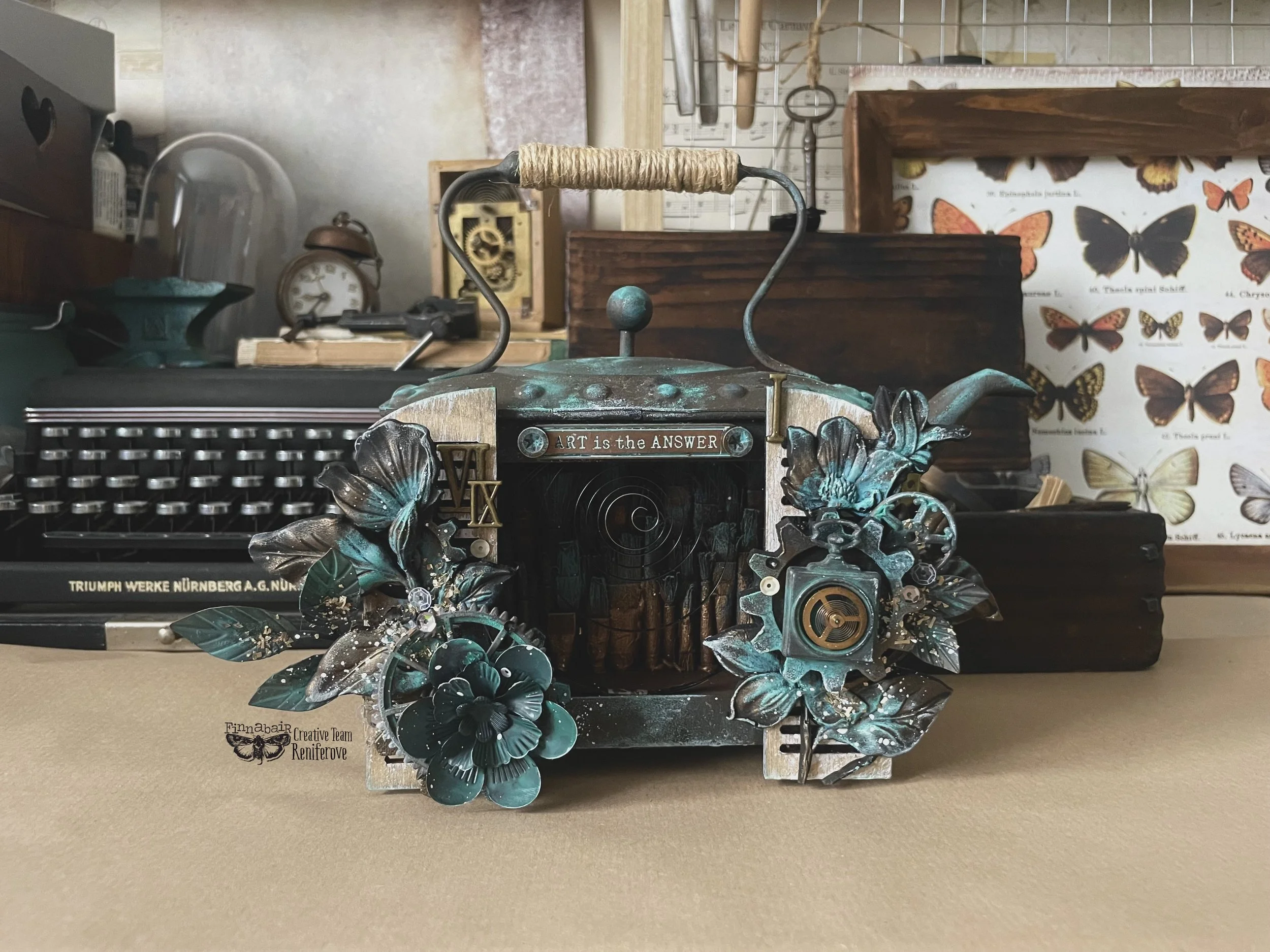 Decorative vintage camera with leaves and flowers, featuring the phrase 'ART is the ANSWER' on top, placed on a table with various vintage items and butterfly illustrations in the background.