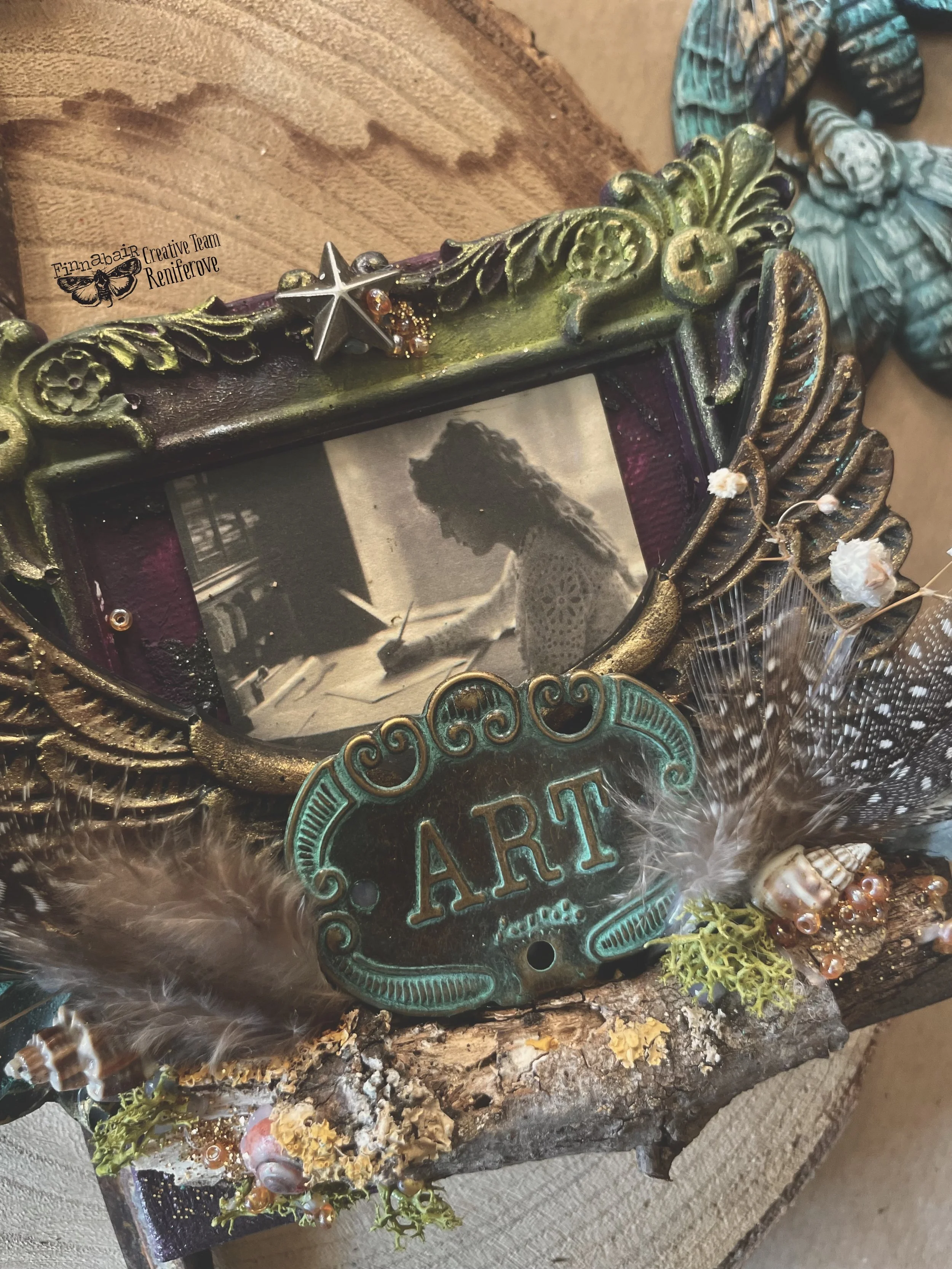 Decorative picture frame with a black-and-white photo of a woman writing, surrounded by art supplies, feathers, shells, moss, and a vintage style sign that says 'ART'.