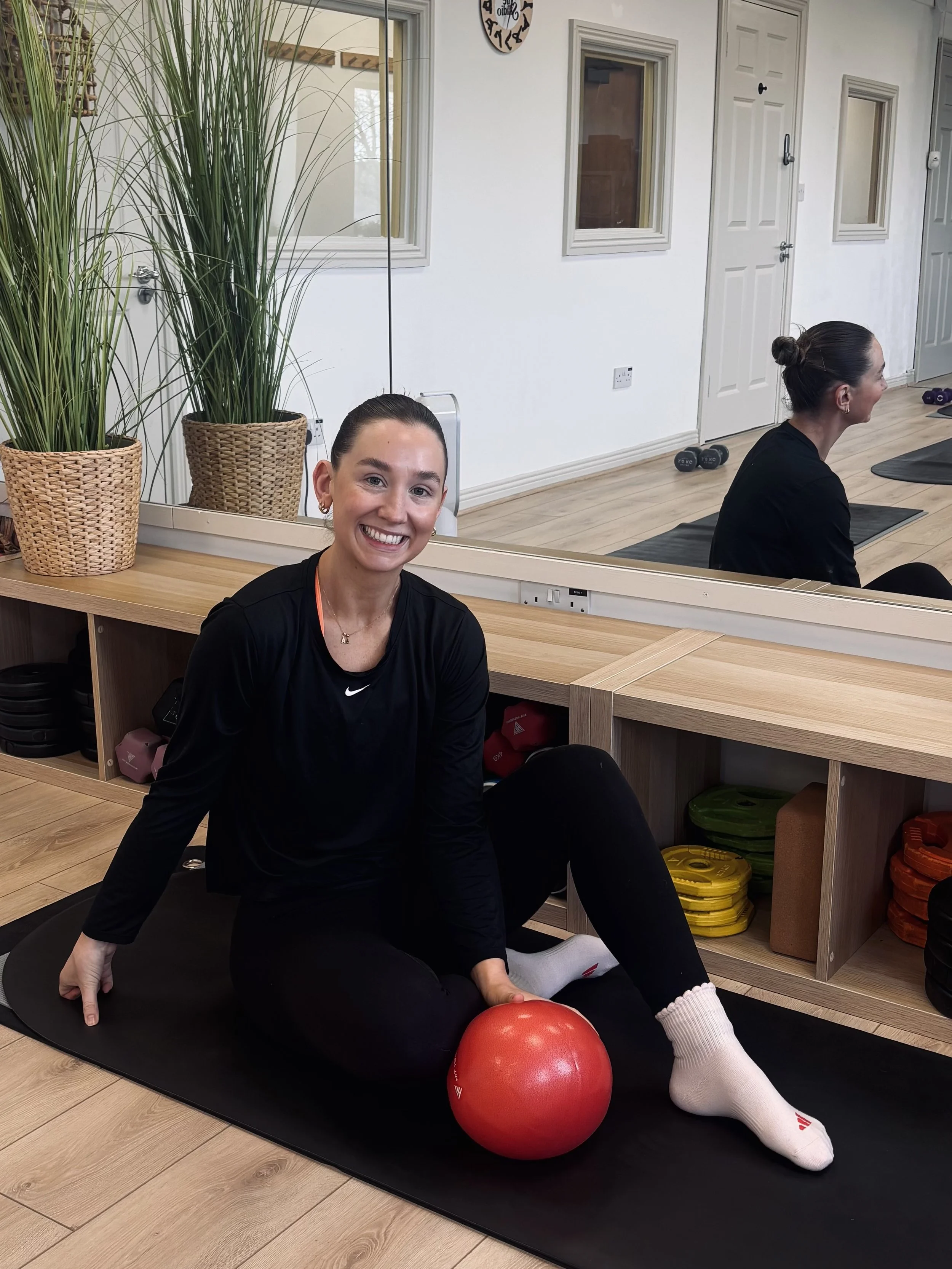 Two women in a gym doing exercises, one sitting on the floor smiling at the camera with a red exercise ball in front of her, and the other in the mirror visible doing a different exercise.