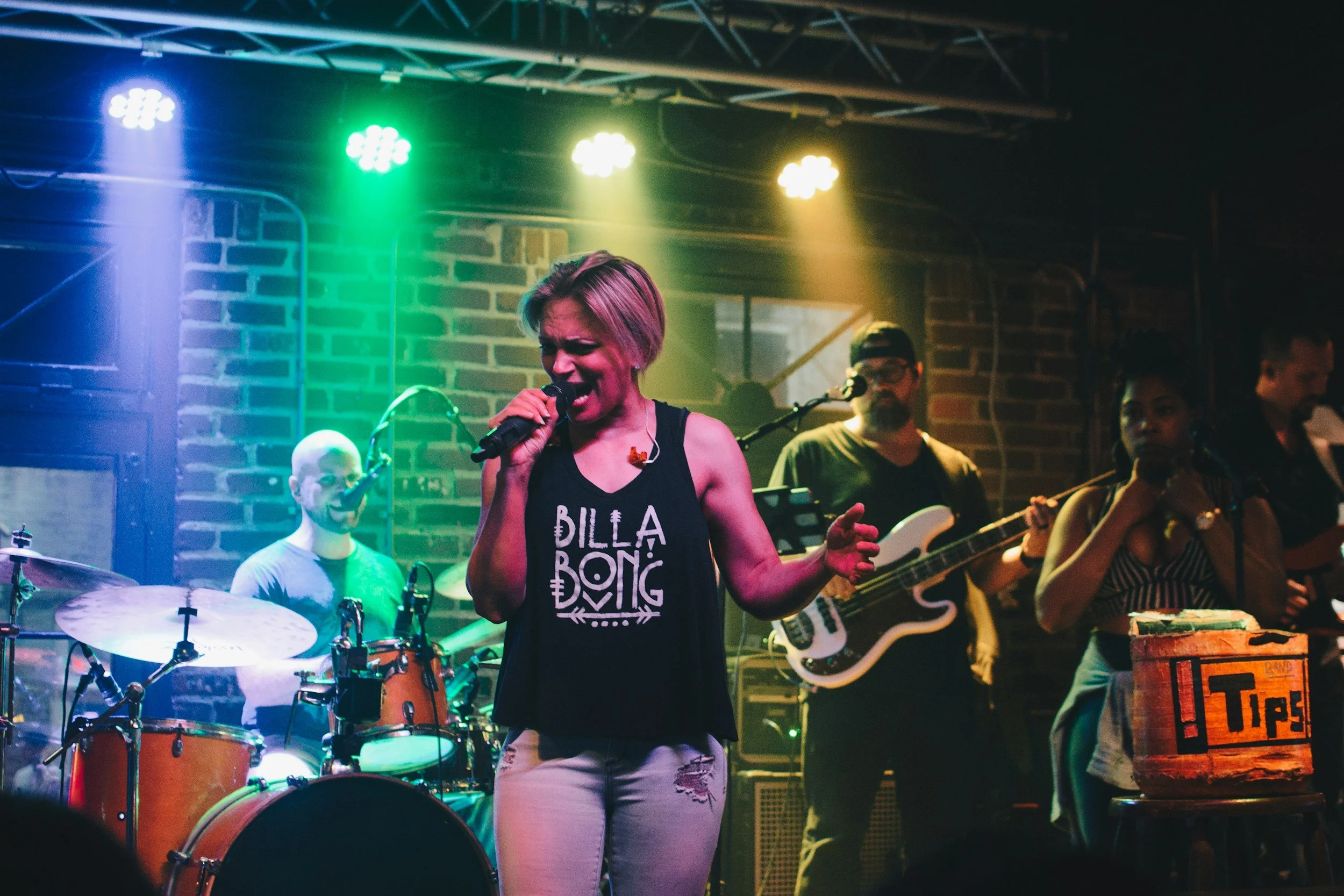 A band performs live on stage with colorful lighting, featuring a female vocalist in a black tank top, with a guitarist, drummer, and keyboardist in the background.