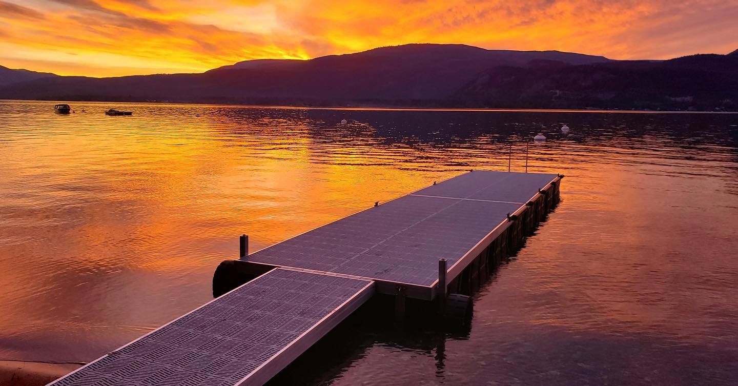 A dock with solar panels on it extends into a calm lake at sunset, with mountains and a colorful sky in the background.