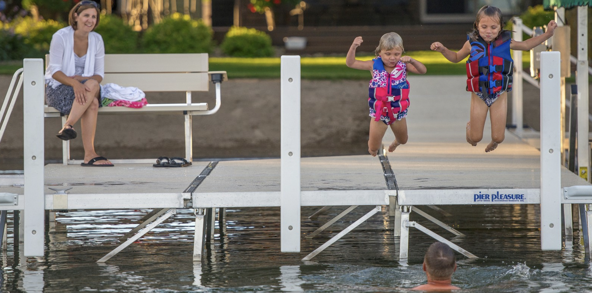 Two young girls in life jackets jumping into the water from a dock while a woman sits on a bench watching and smiling, with the sun setting in the background.