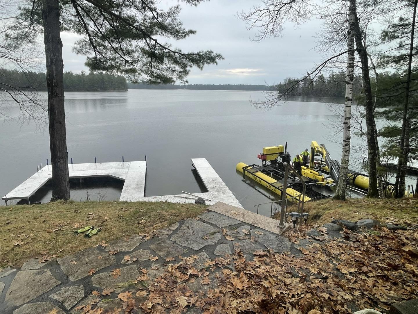 A lakeside scene with a dock under construction and a hovercraft with workers on the water, surrounded by trees and fallen autumn leaves.
