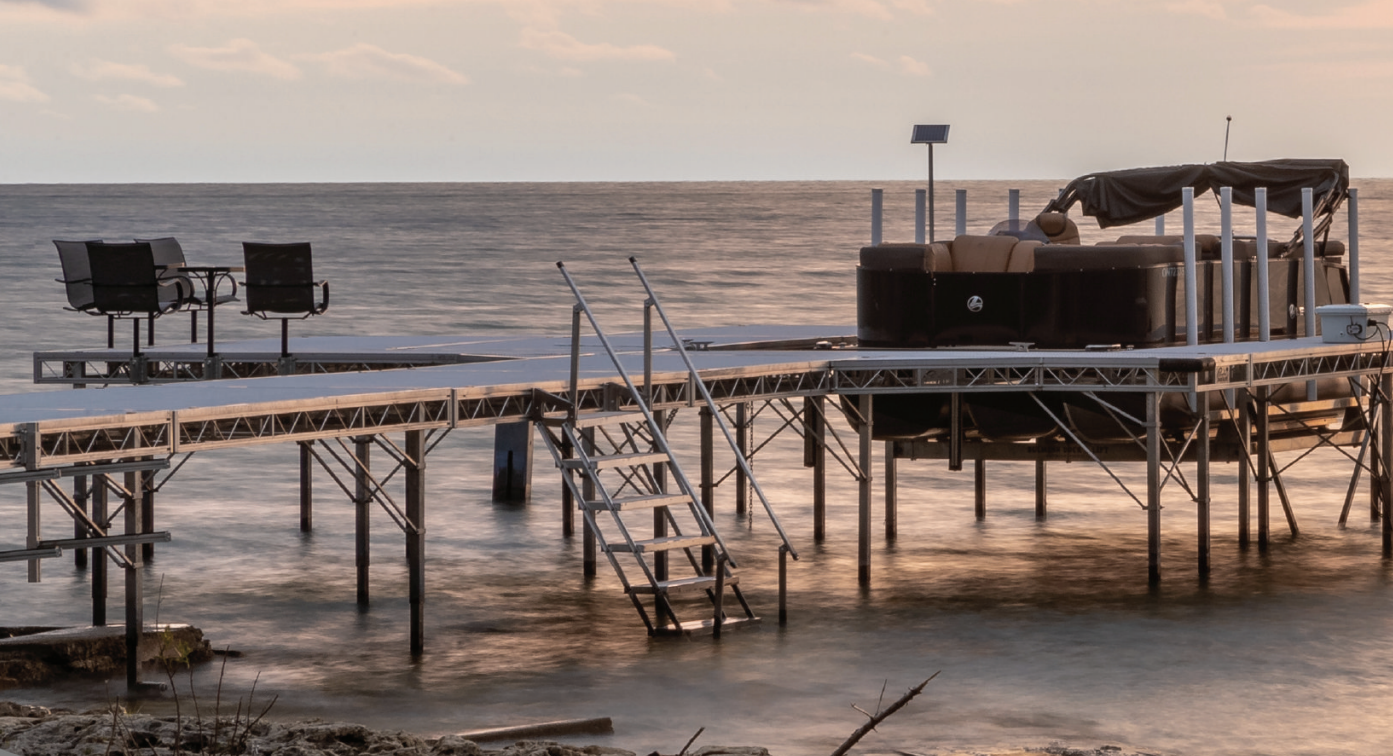 A wooden pier extending into the ocean during sunset with two chairs on the pier, solar panels, and a hot tub on a deck, with calm water and a partly cloudy sky.