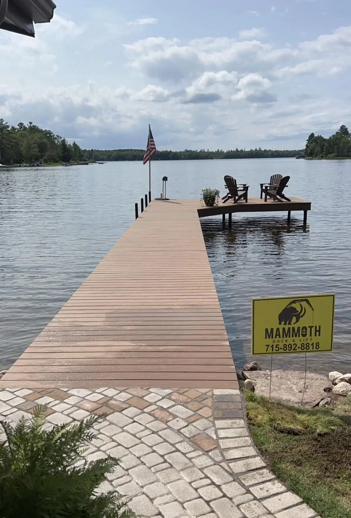 A wooden dock extending into a calm lake with Adirondack chairs at the end. An American flag is mounted on the dock, and there is a potted plant nearby. The sky is partly cloudy, and there is a sign on the shore that says "MAMMOTH Dock & Lift" with a