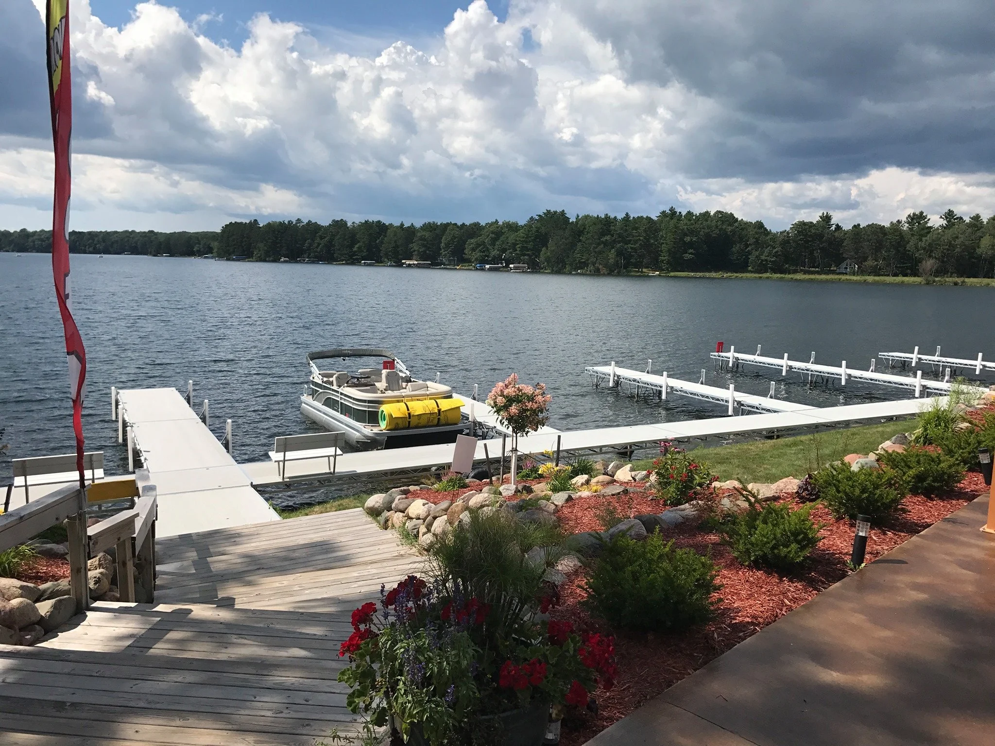 A lakeside scene with a pier, a pontoon boat, and a dock, with greenery, flowers, and rocks along the shore under a cloudy sky.