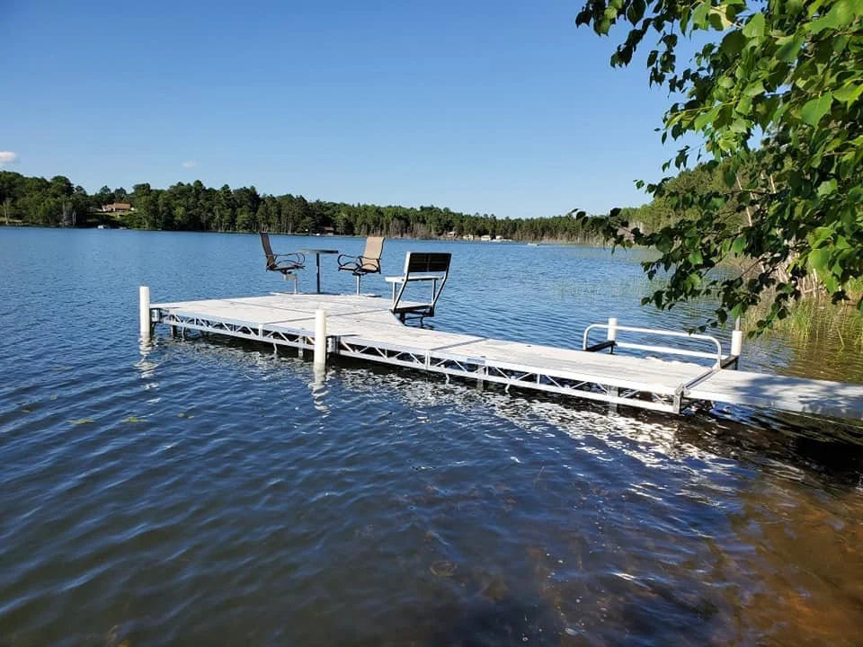 Floating dock with three chairs on a lake, surrounded by trees under a clear blue sky.