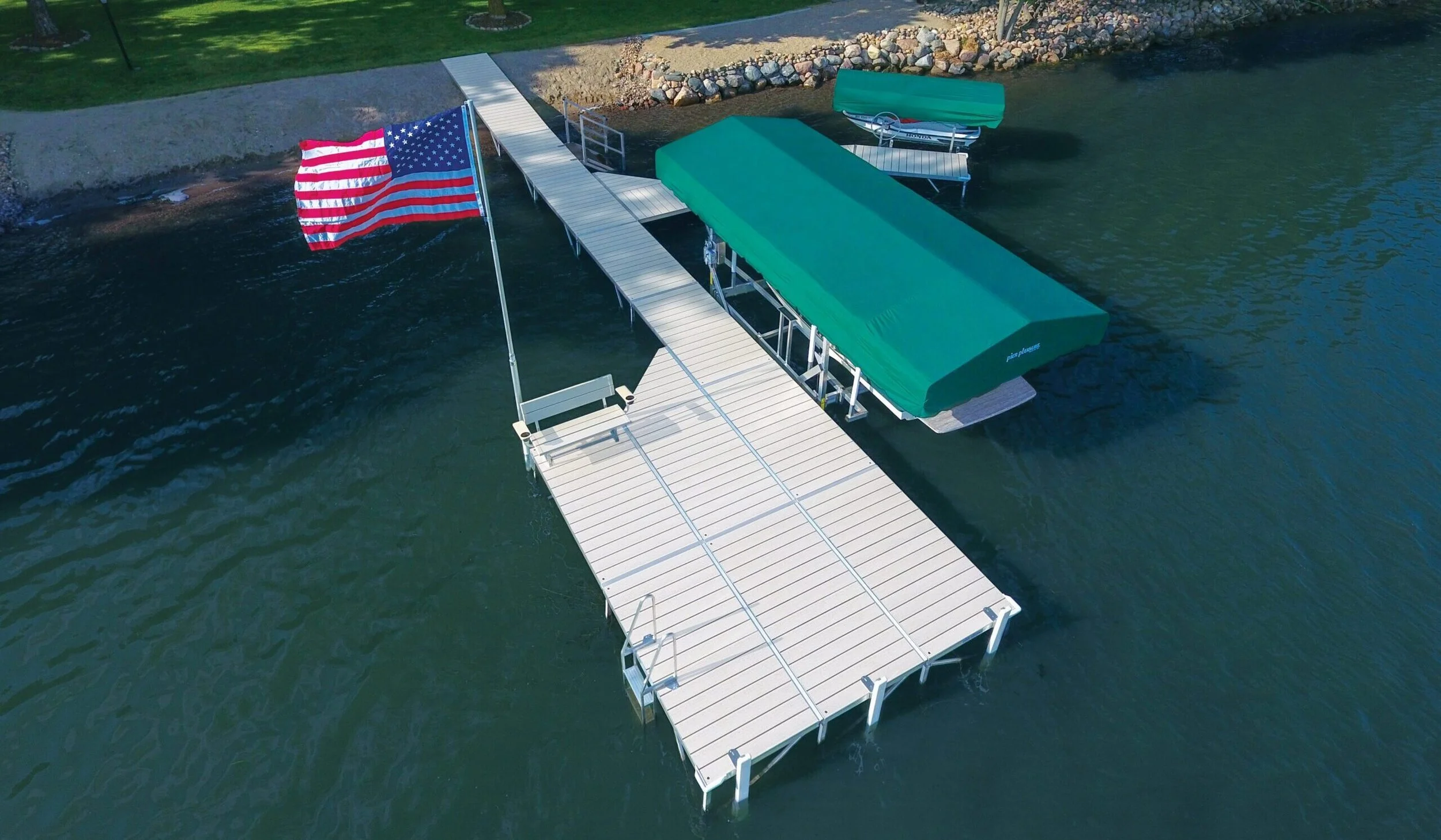 Aerial view of a dock with a staircase leading to a boat with green covers, a flagpole with an American flag, and a small boat in the water near the dock.