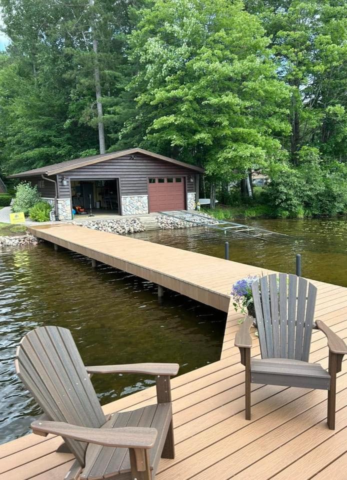 A lakeside scene with a wooden dock leading to a boathouse. Two Adirondack chairs are positioned on the dock, one facing the water and the other facing slightly to the side. There are trees and greenery surrounding the boathouse and along the shoreli