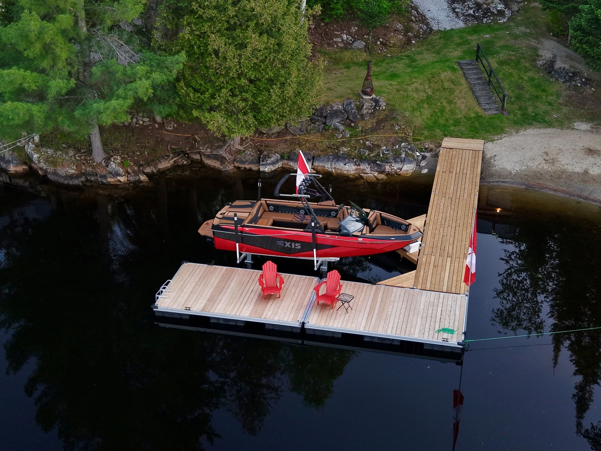 Aerial view of a red and black boat docked at a wooden pier on a calm lake, with two red chairs on the pier and green trees along the shoreline.