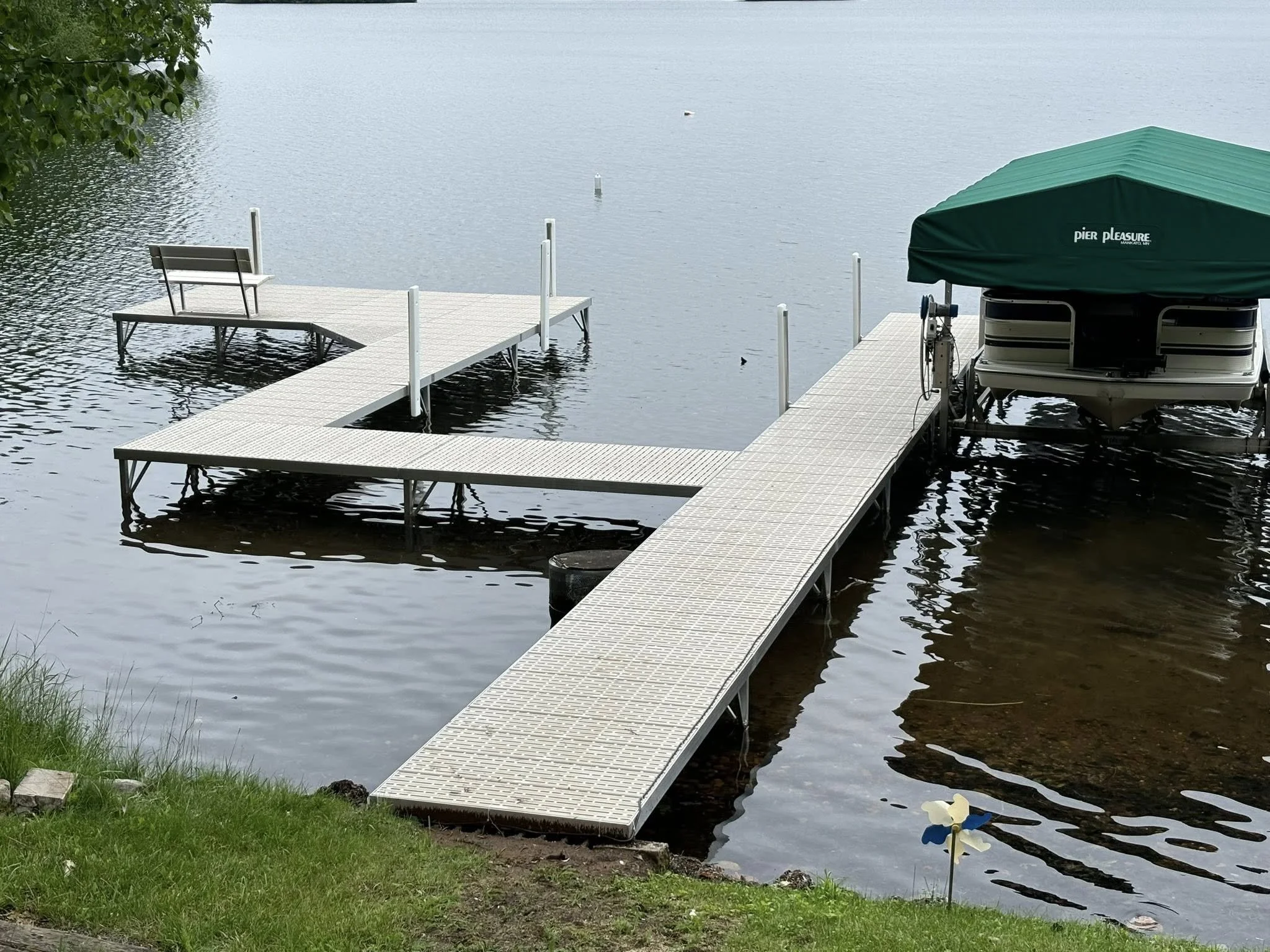 A lake dock with metal ramp leading to a boat with a green canopy, a small bench at the end of the dock, bordered by green grass and a yellow and white flower at the shoreline.