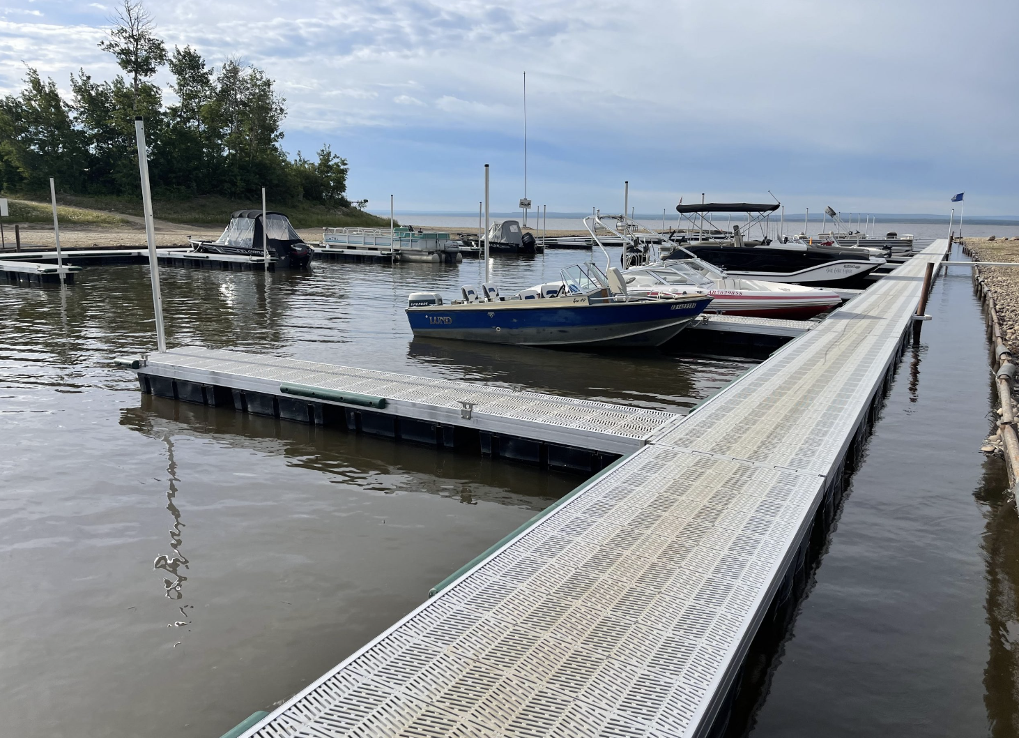 A marina with boats docked, a floating dock extending into the water, and a shoreline with trees in the background under a cloudy sky.