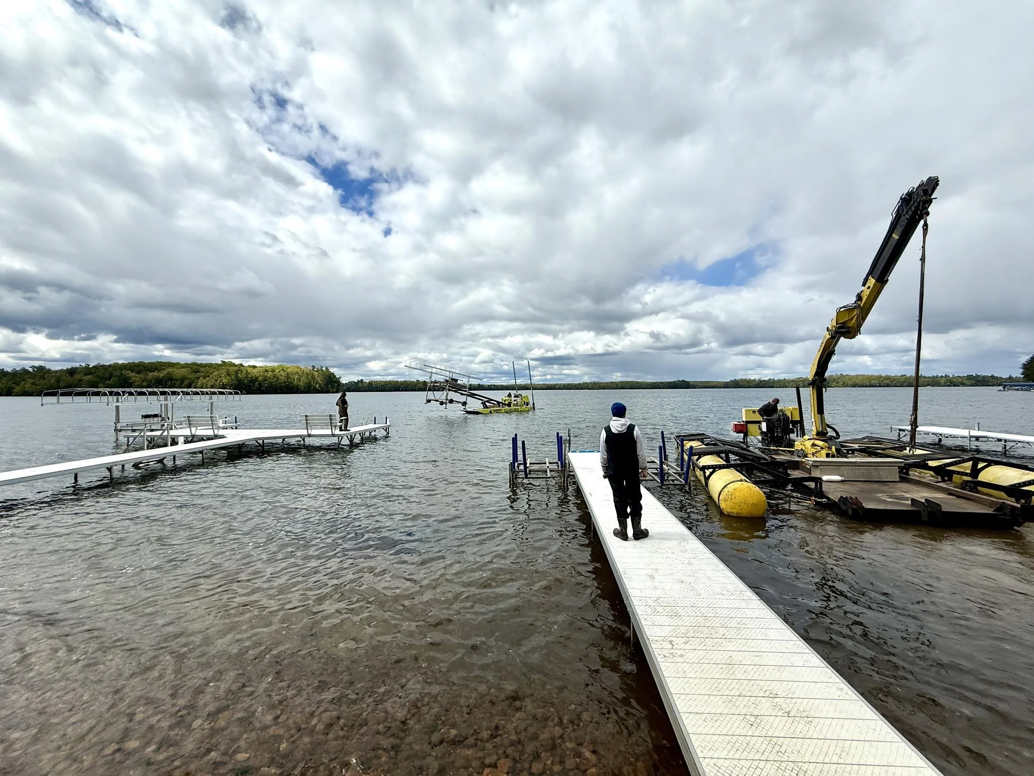 Overcast sky with scattered clouds over a large body of water. A person stands on a dock, watching a boat being assembled or repaired with a crane on a floating platform. Another person stands on a different dock in the distance. Trees line the horiz