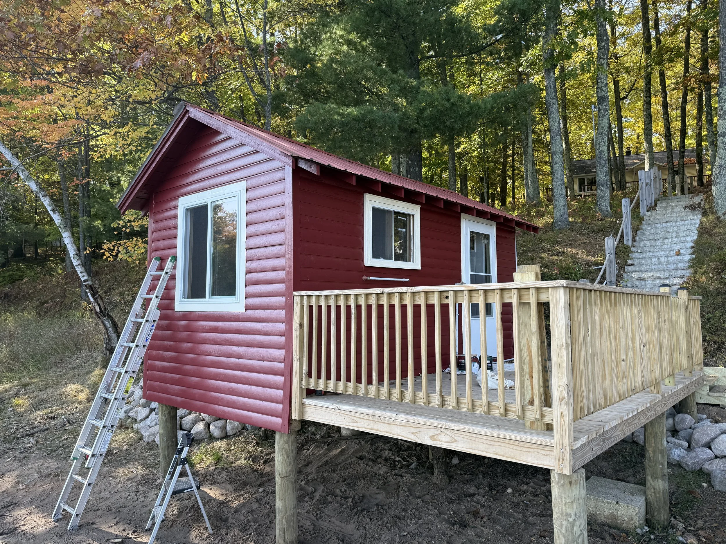 A small red wooden cabin with a deck in the woods during autumn, with a ladder leaning against one side and a set of stone stairs leading up the hill behind it.