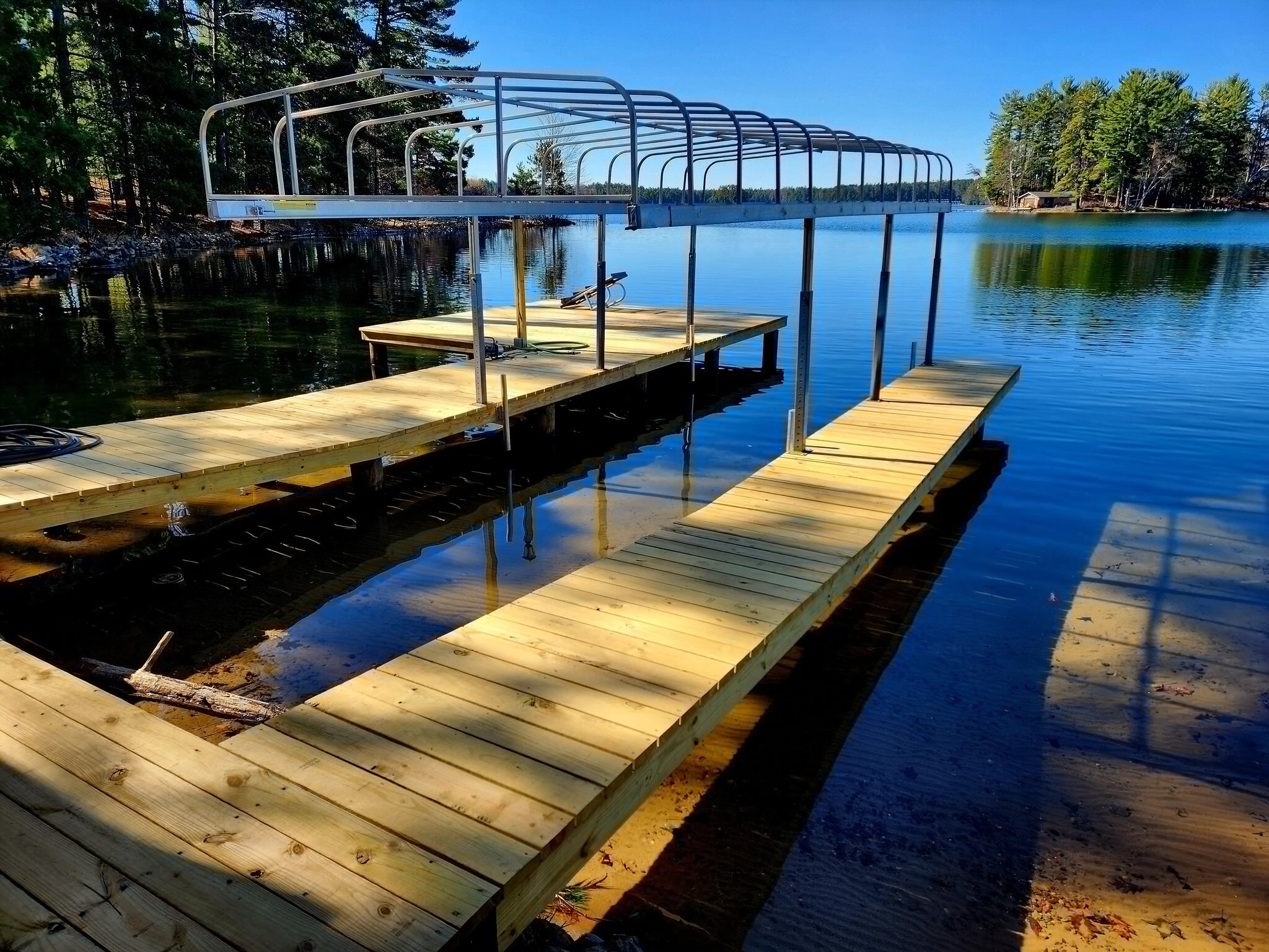 Wooden dock with metal boat lift extending over calm lake surrounded by trees under a clear blue sky.