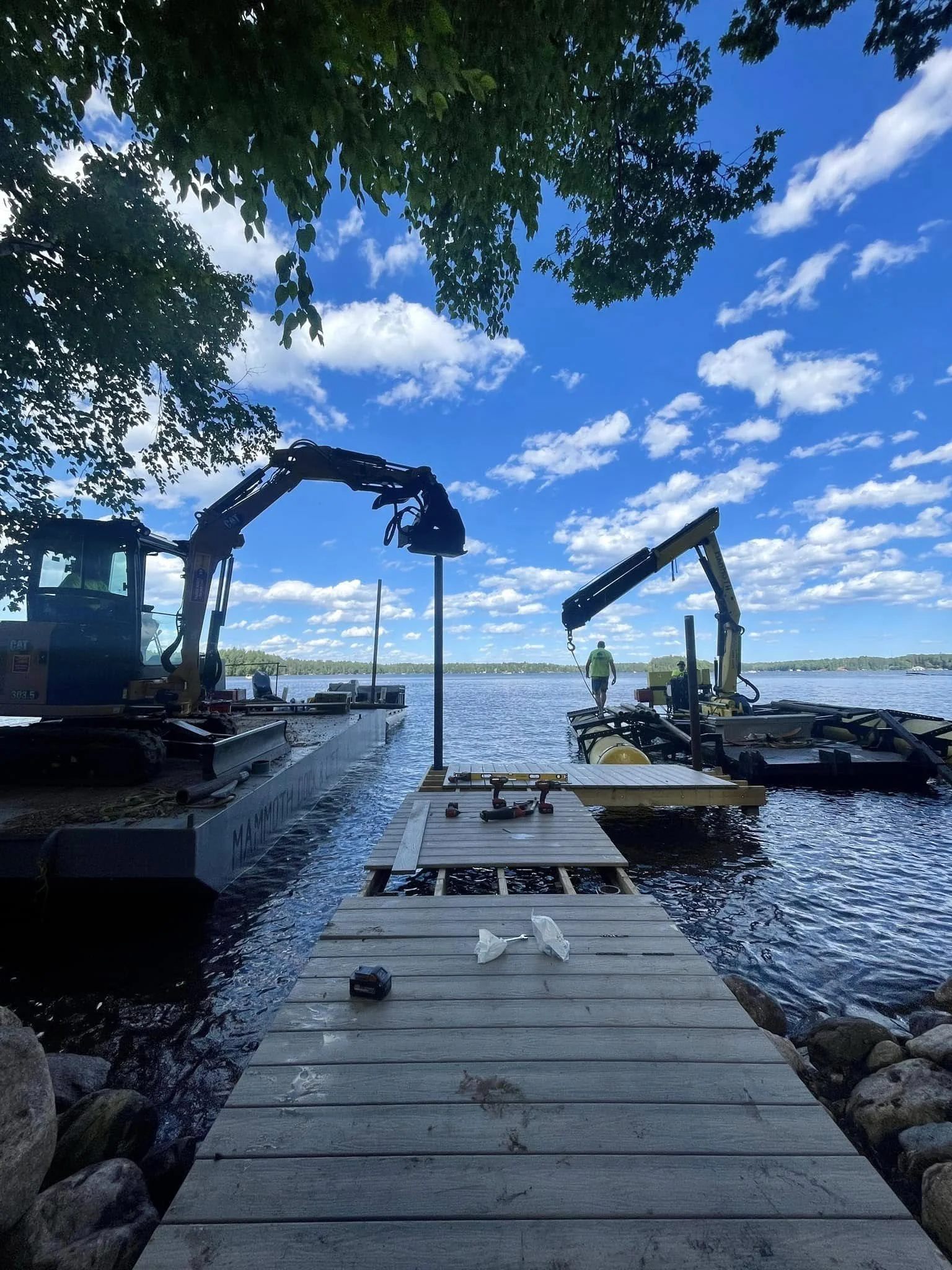 Construction equipment on a dock by the water with two cranes, a worker, and a partly cloudy sky.
