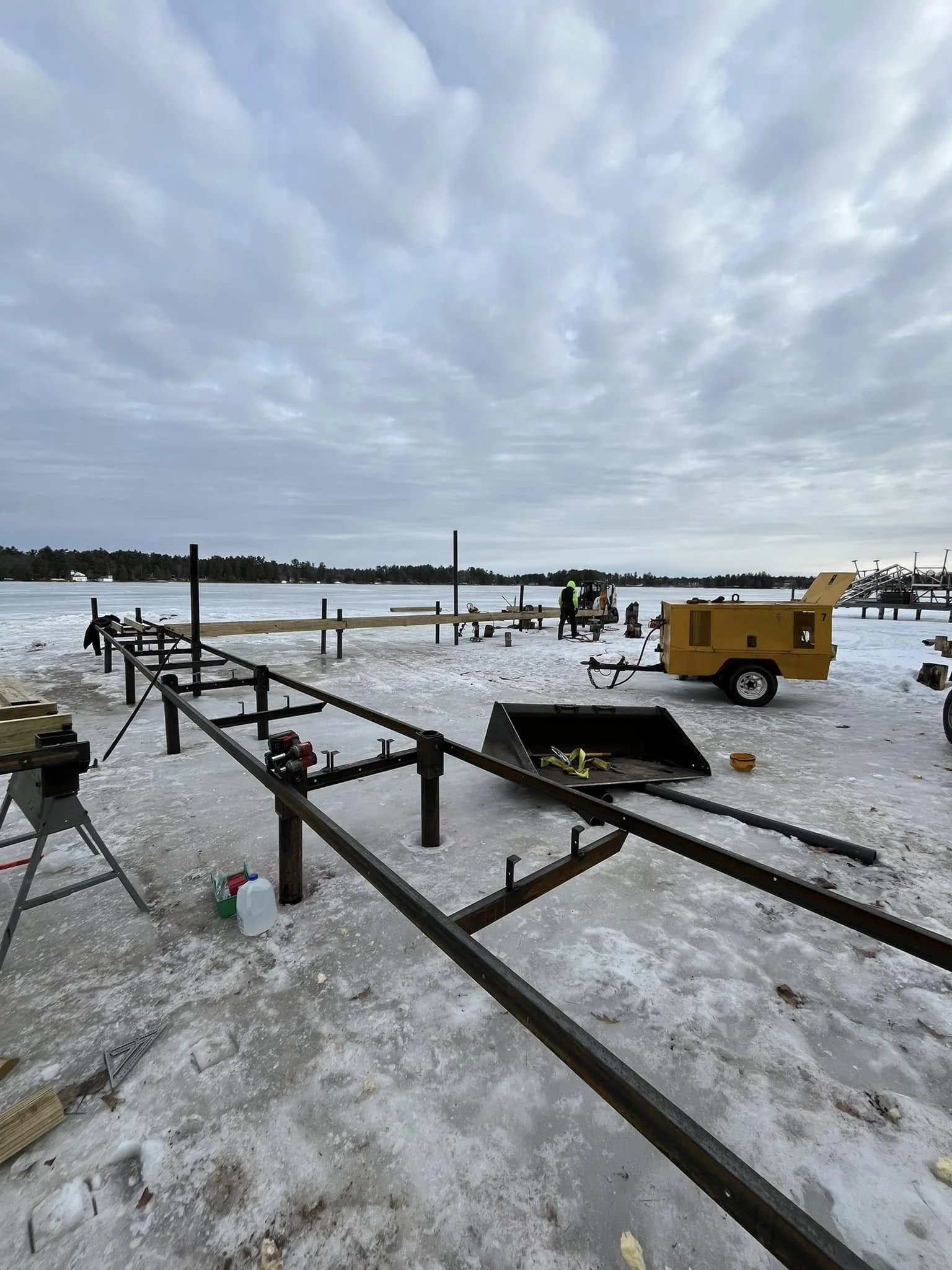 Construction site by a frozen lake with workers, yellow equipment, and metal framework for a dock, under a cloudy sky.