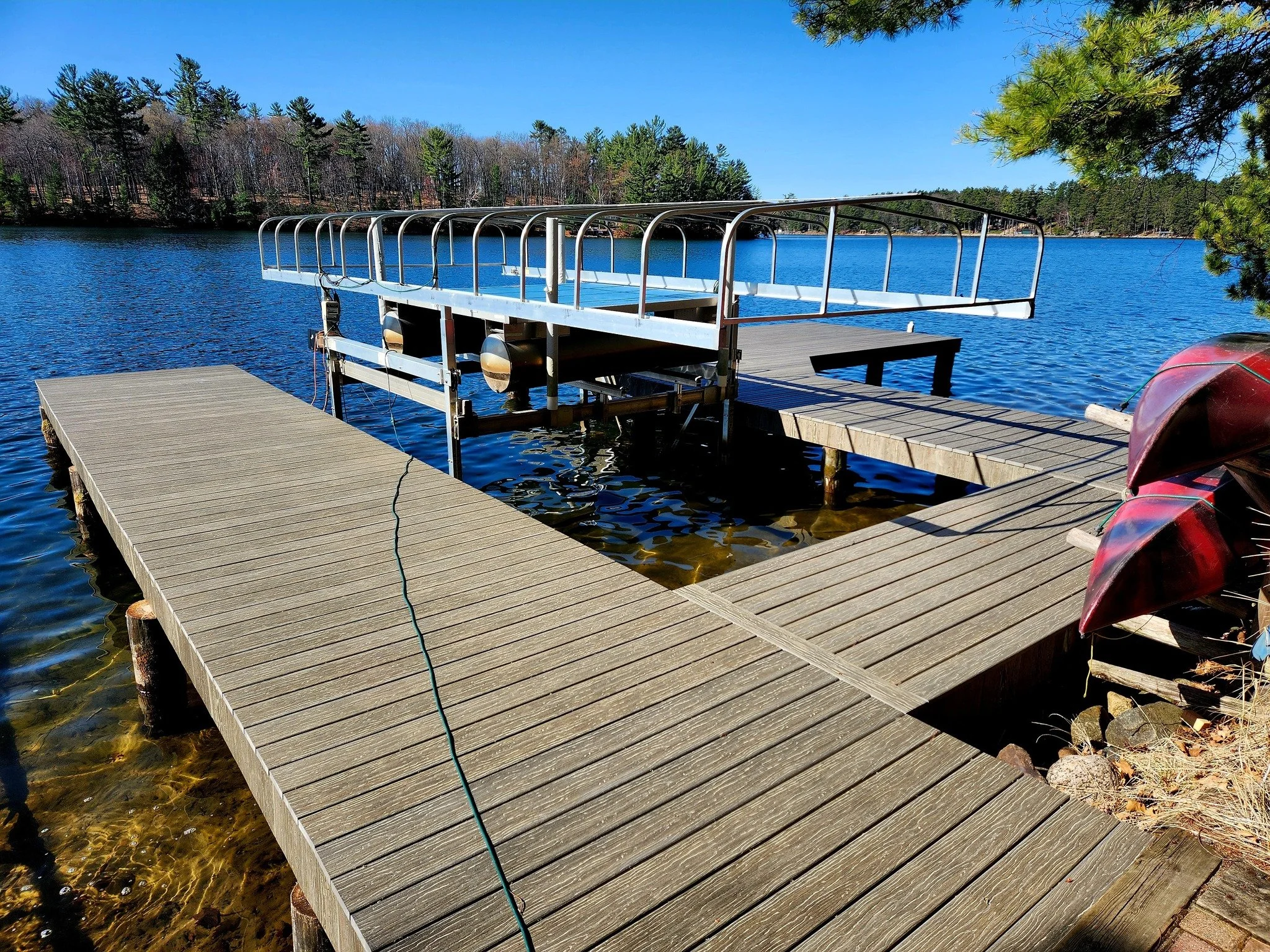 Wooden dock extending into a lake with a boat lift structure at the end, surrounded by water and trees in the background, clear blue sky above.