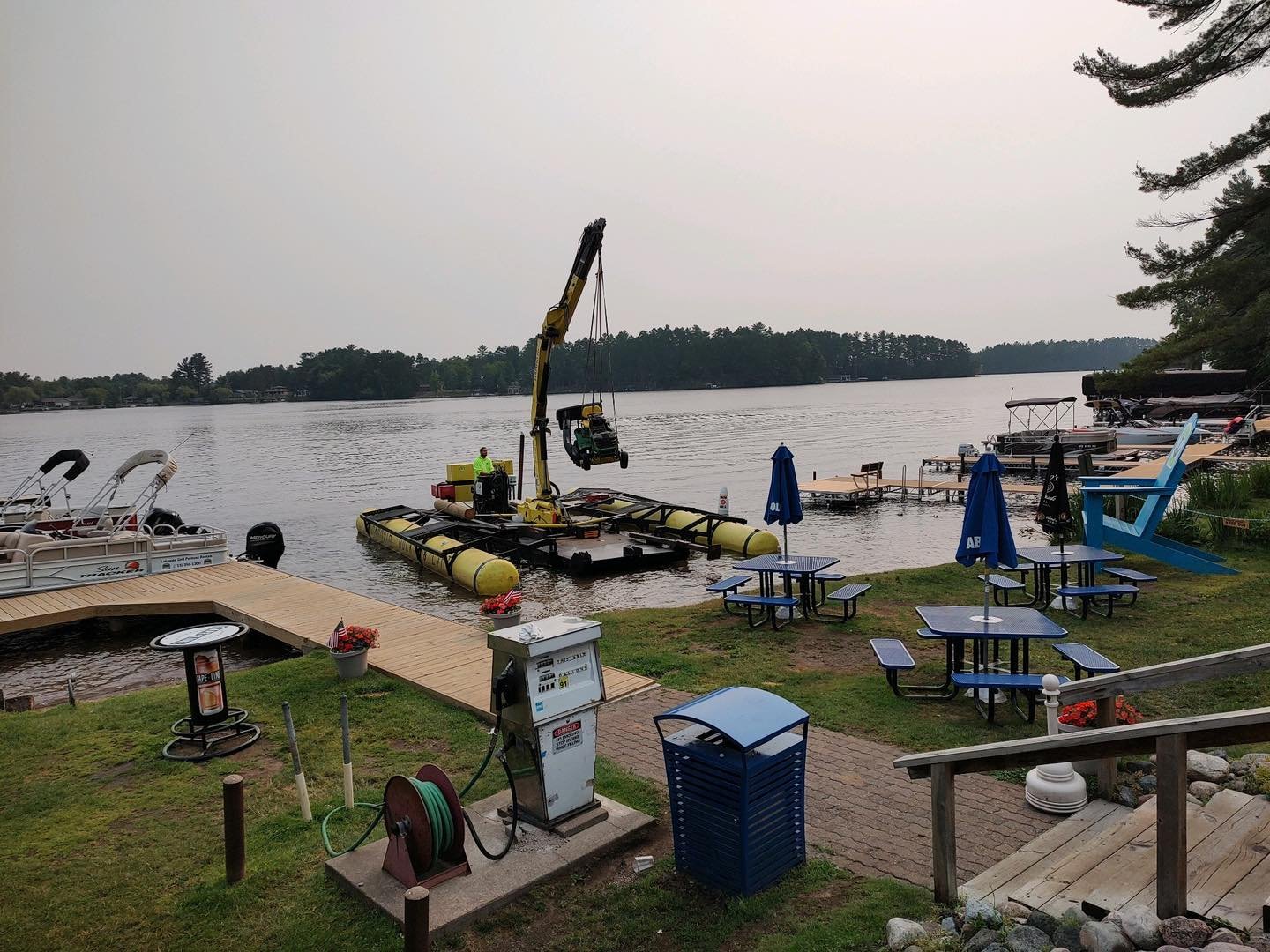 A lakeside scene with docked boats, a floating crane, empty picnic tables with umbrellas, and a gravel walkway, under a cloudy sky.