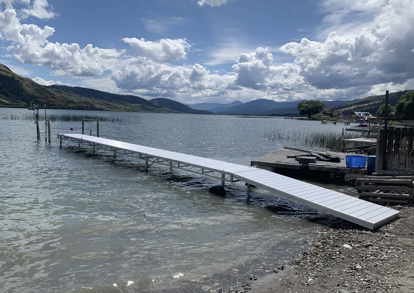 A lakeside scene with a dock extending into the water, surrounded by mountains, cloudy sky, and houses along the shore.
