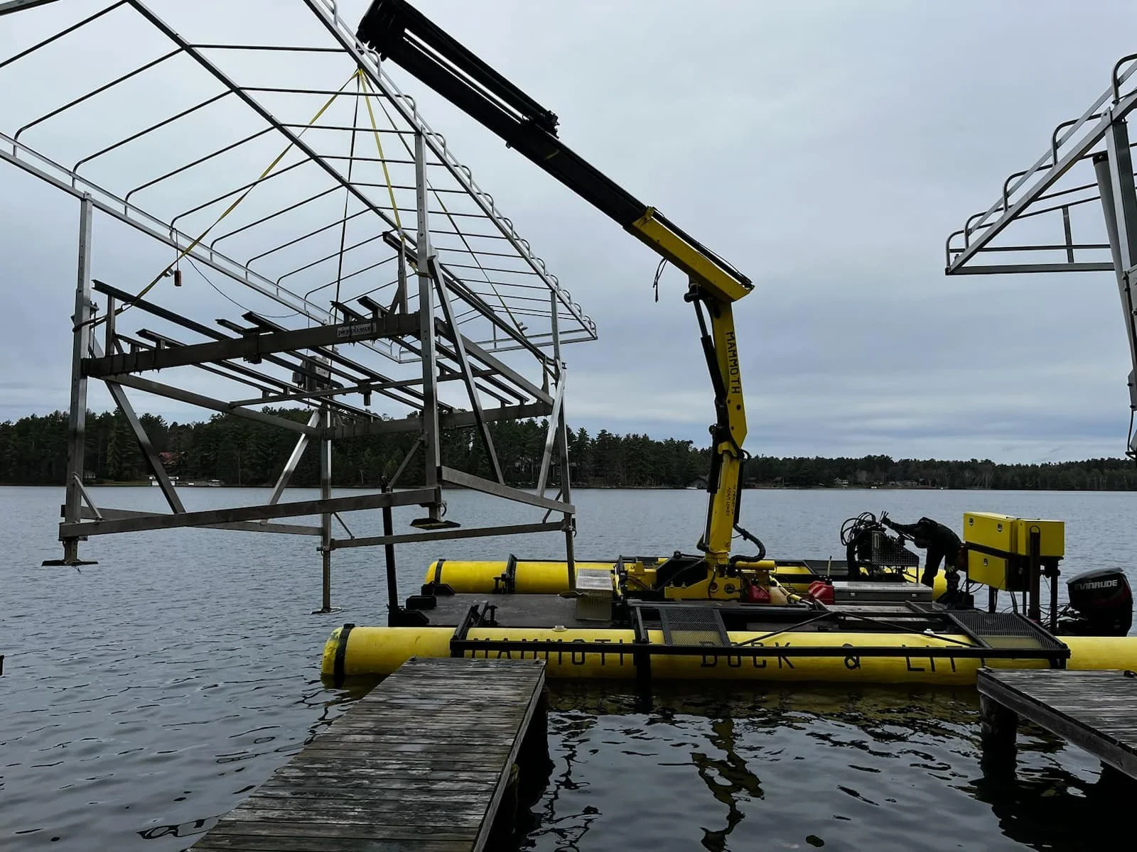 A yellow amphibious drone equipped with a crane lifting a metal structure over a lake, with a dock in the foreground and a tree-lined shoreline in the background under a cloudy sky.
