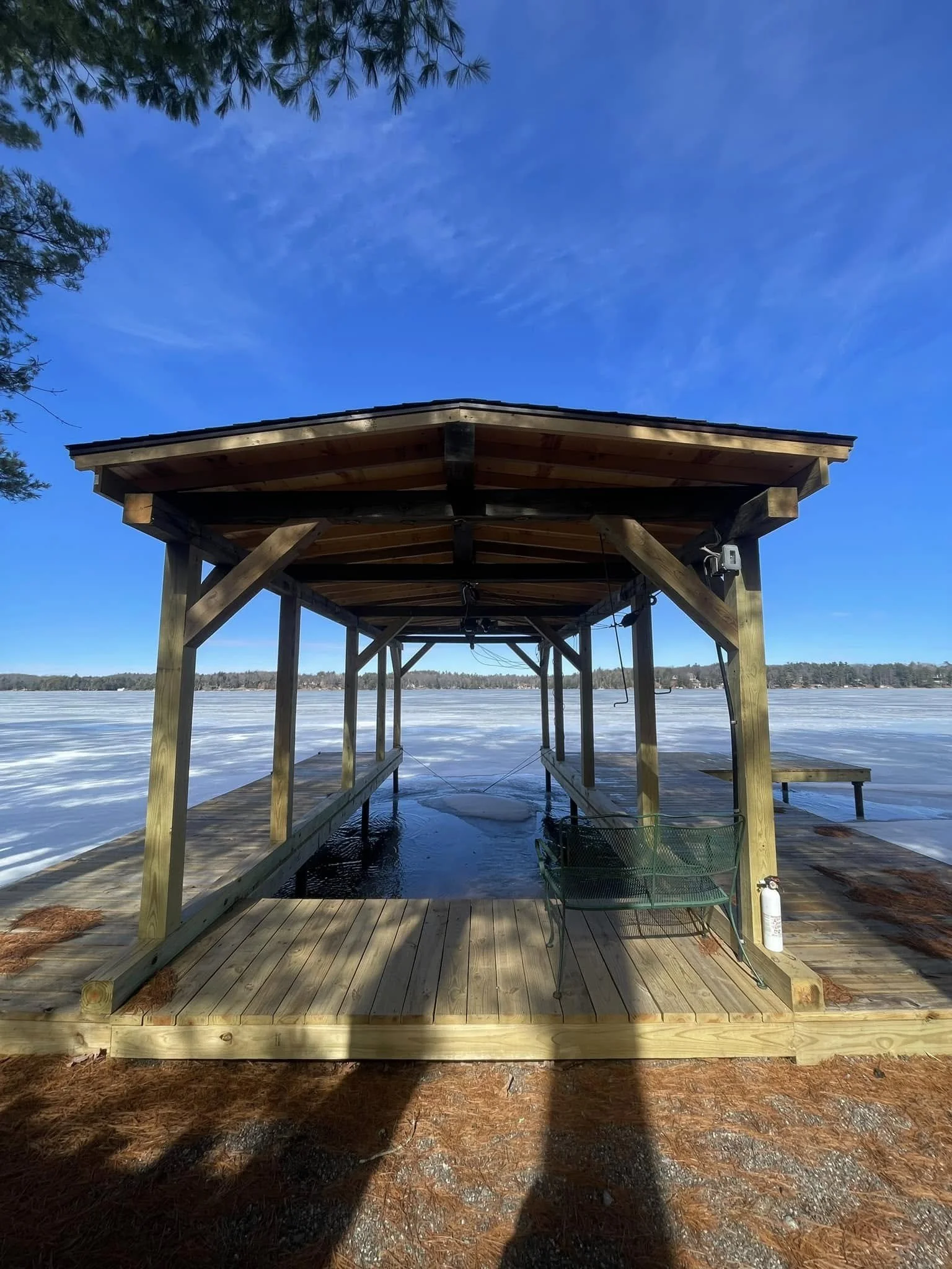A wooden dock with shelter extending over a frozen lake, under a bright blue sky with some clouds, surrounded by trees.