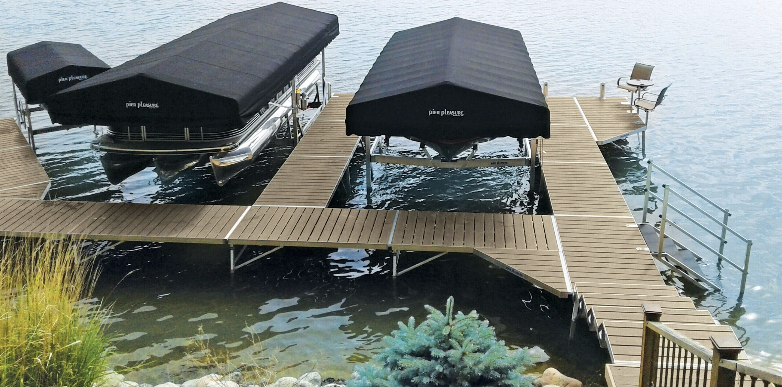 A wooden dock extending into a lake with two boat lifts covered by black canopies, each holding a pontoon boat. There is a small seating area with two chairs on the dock, and the water is calm.