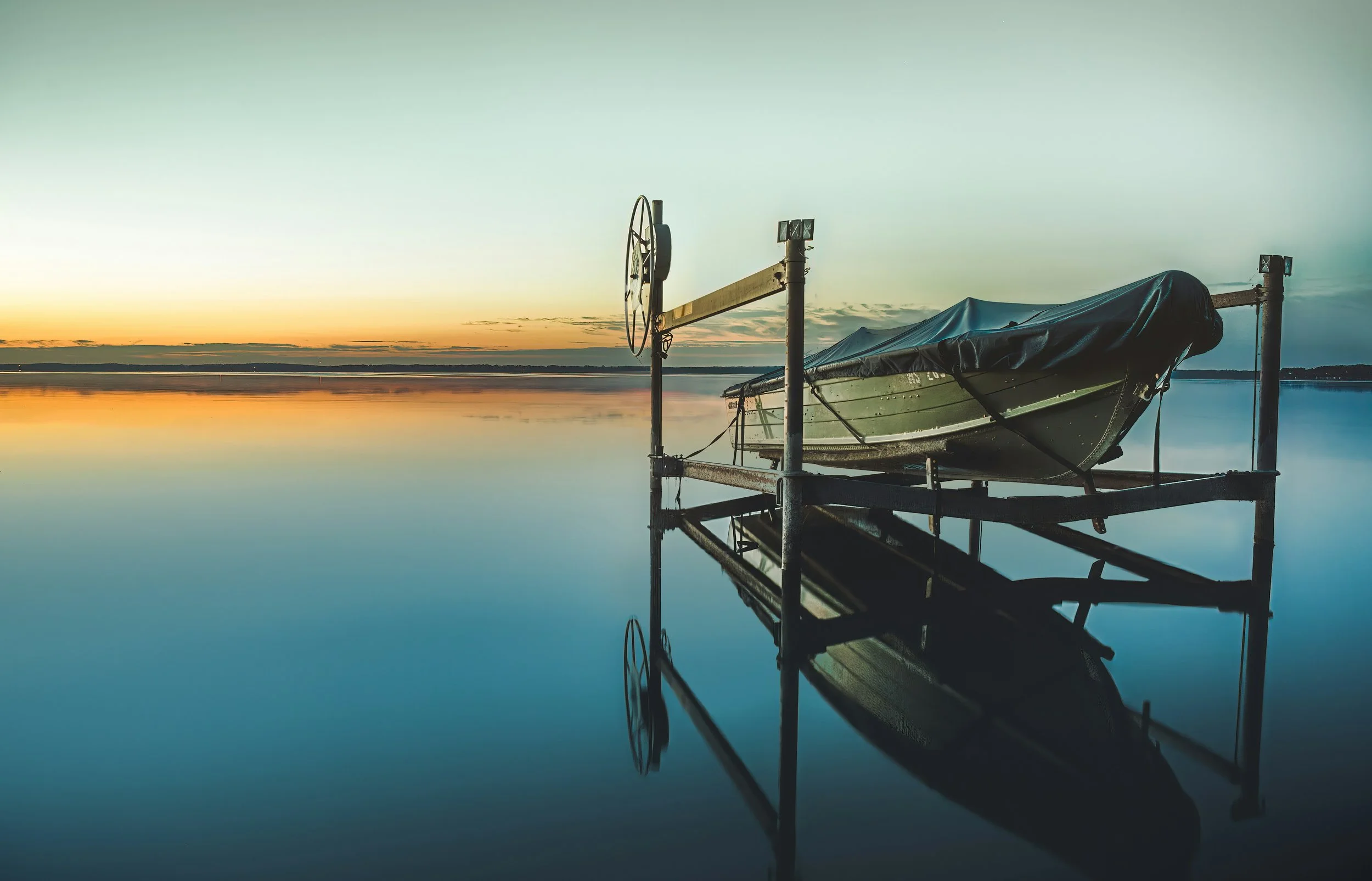 A boat covered with a tarp resting on a dock by calm water during sunset or sunrise, with its reflection visible in the water.