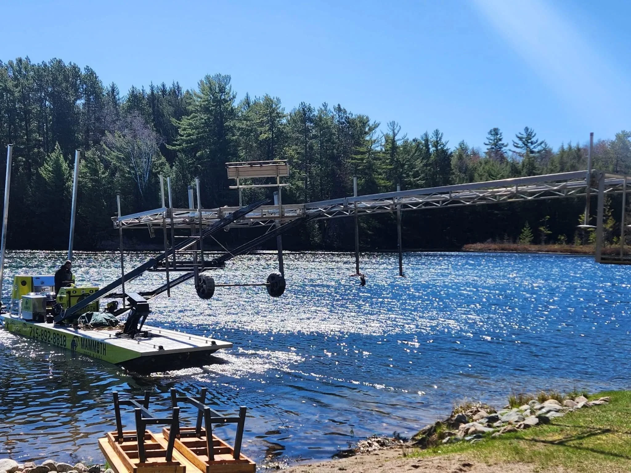 A flood barrier being installed across a body of water near a forested shoreline, with a construction platform and machinery, under a clear sunny sky.