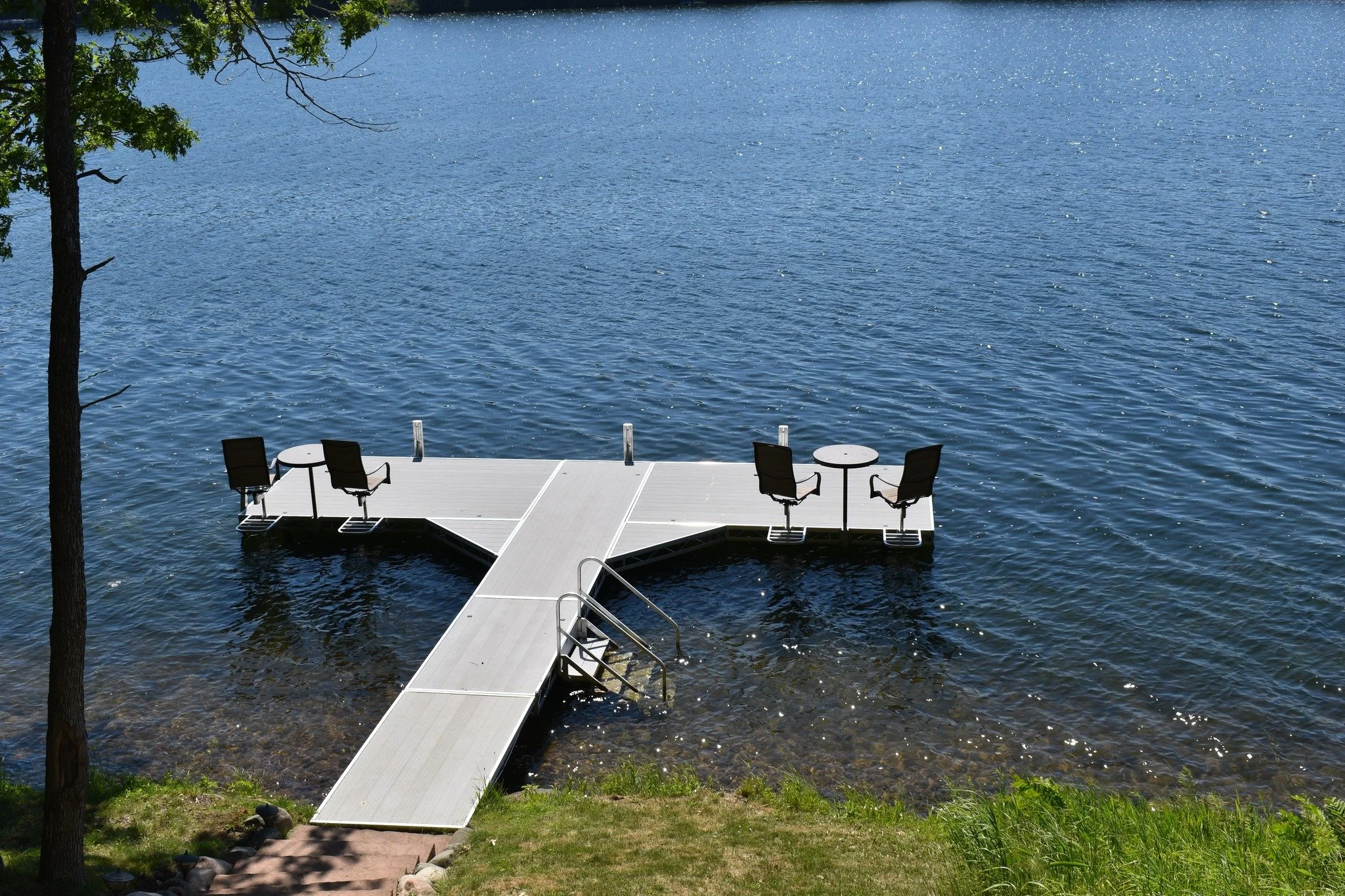 A wooden dock extends into a calm lake, with two chairs and small tables on the dock, surrounded by grassy shoreline and trees.
