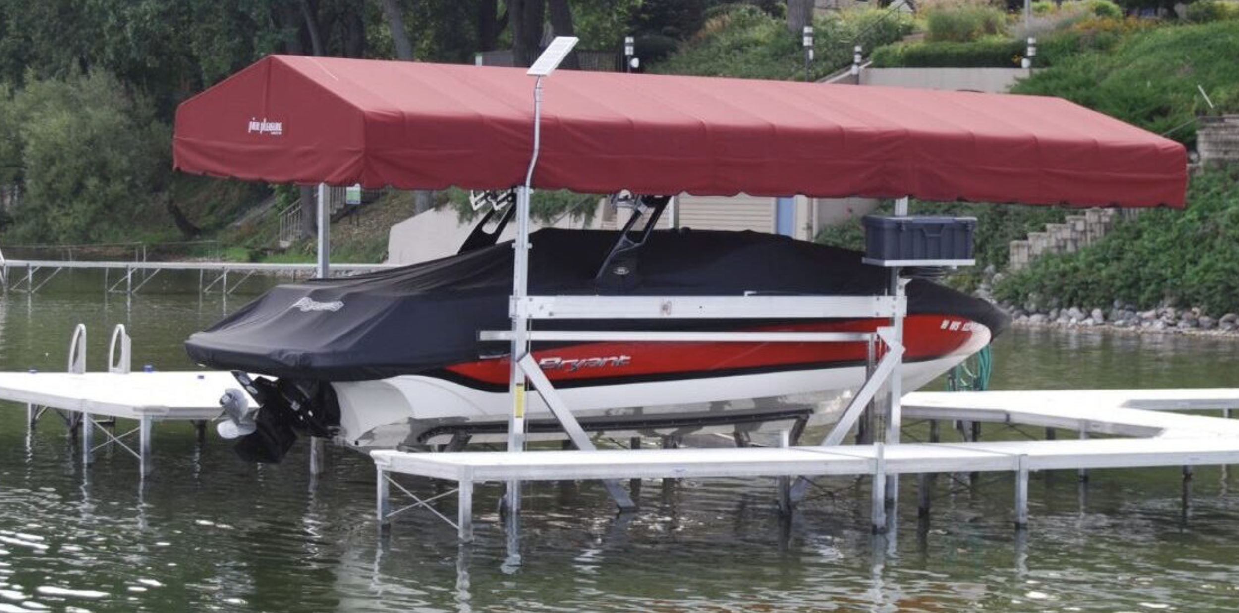 A black and red speedboat docked on a floating water platform with a large red canopy and a white walkway surrounding it, near a river with trees and stairs in the background.